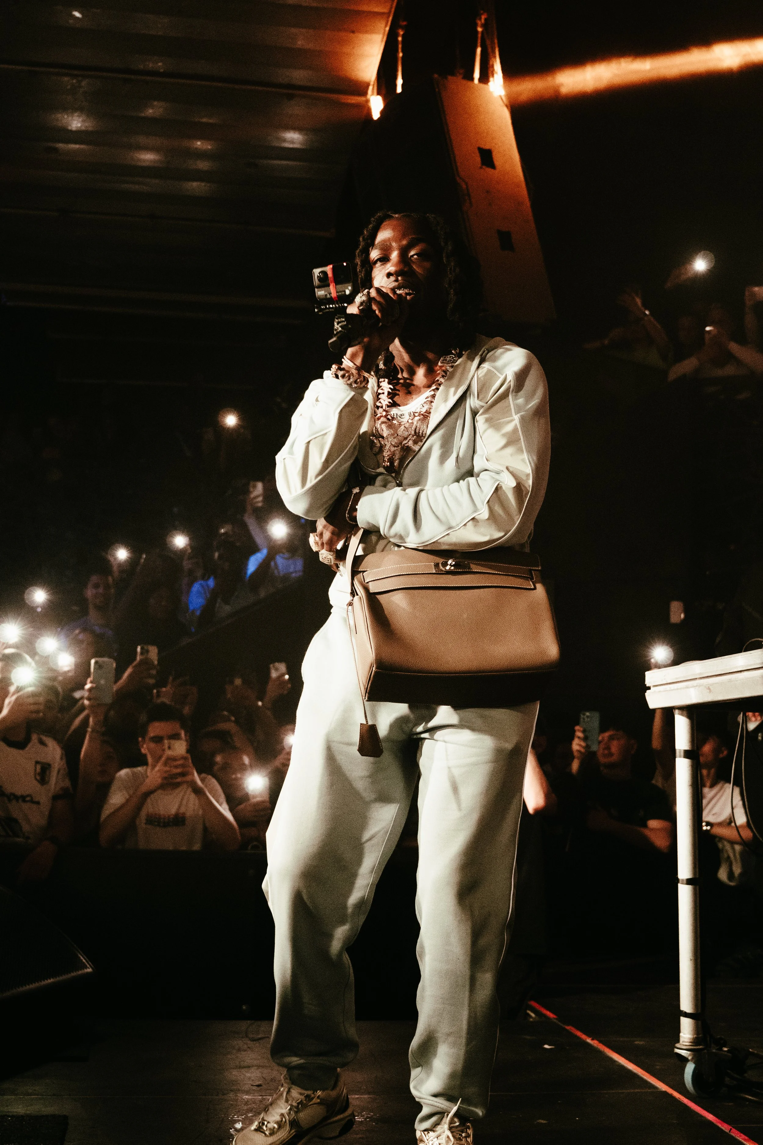 Femme parlant dans un microphone sur scène lors d'un concert, entourée de spectateurs avec des lumières de téléphone portables allumées.