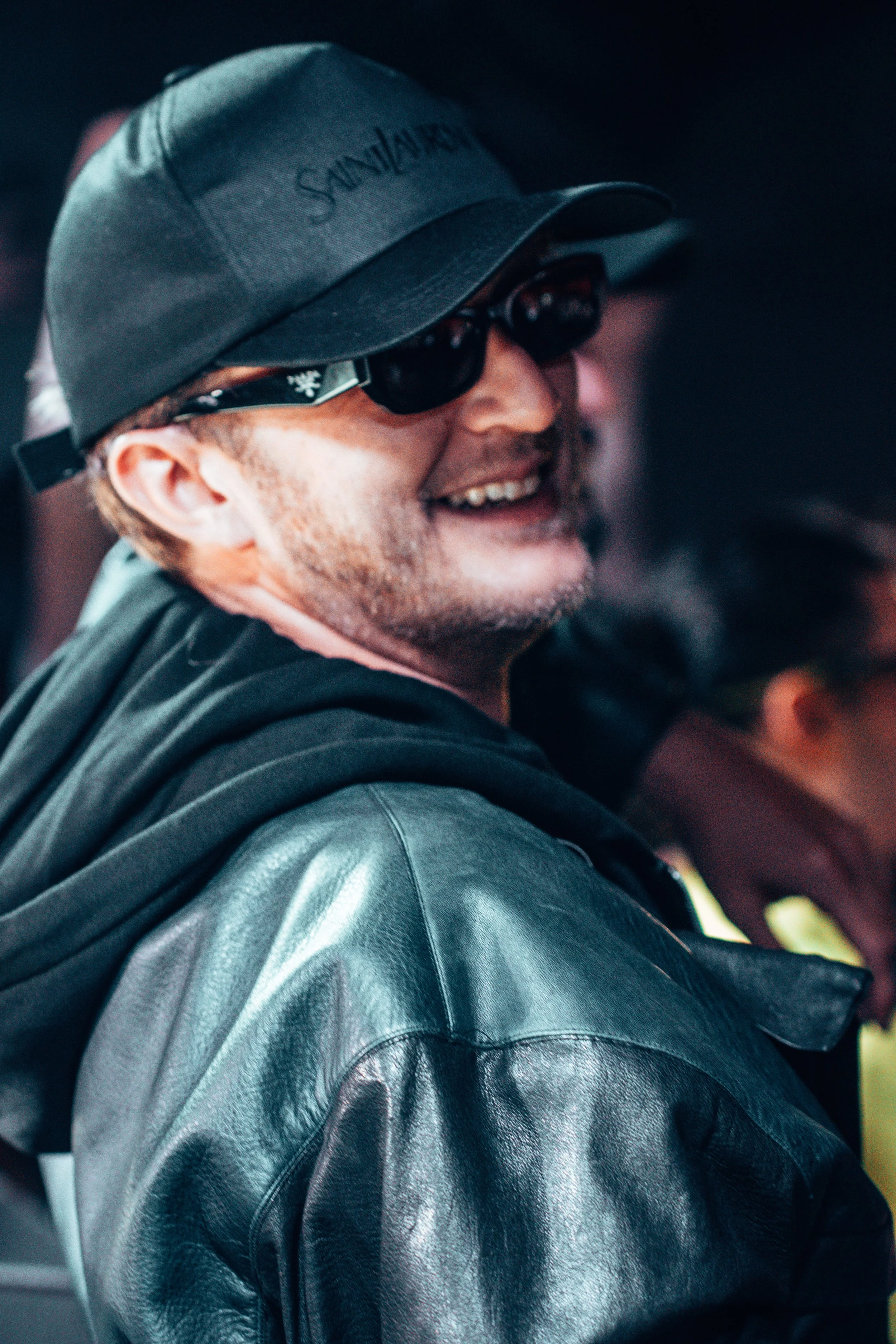Un homme souriant portant une casquette, des lunettes de soleil et une veste en cuir.