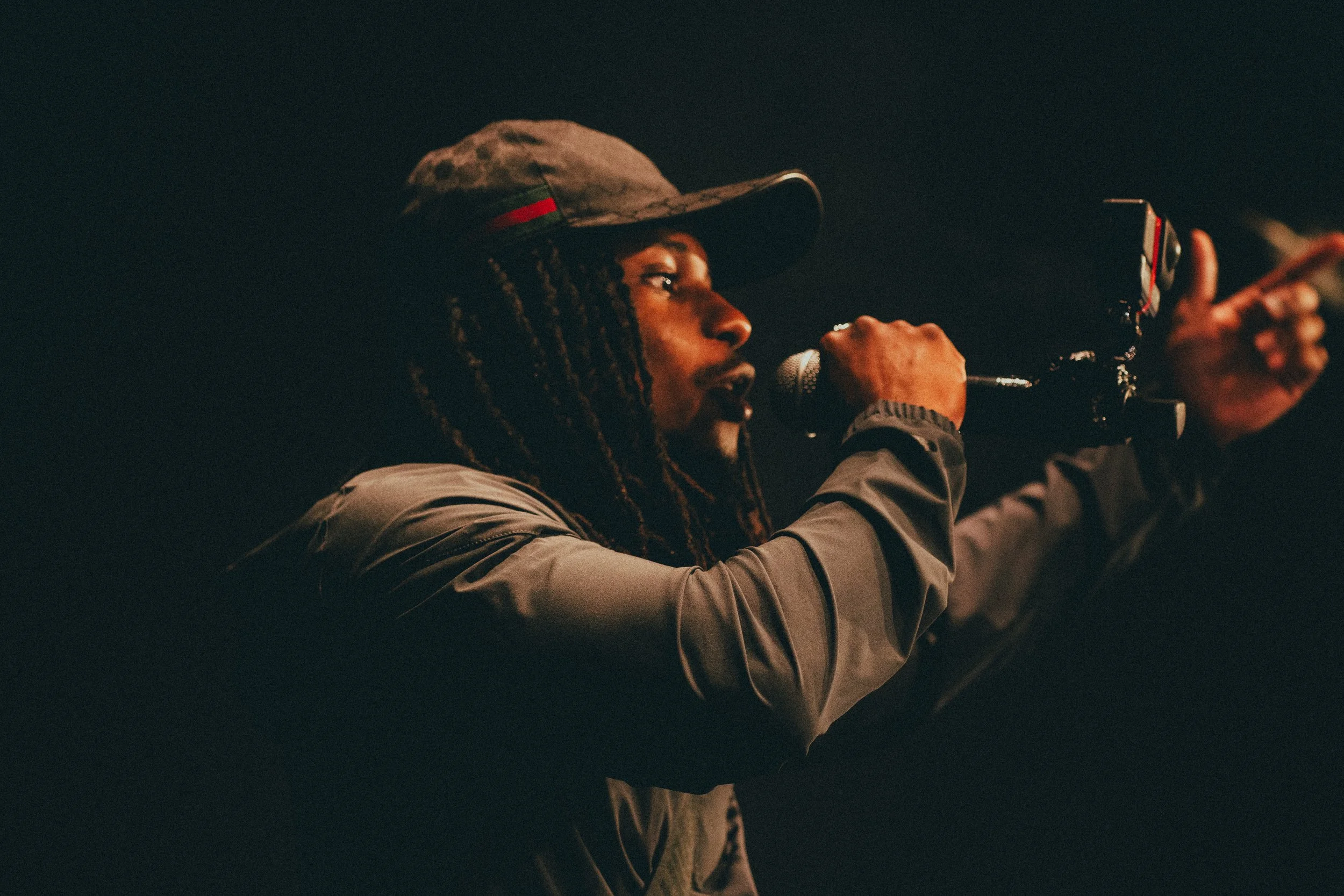 Une personne portant un chapeau de baseball, tenant un microphone et pointant avec un pistolet factice.