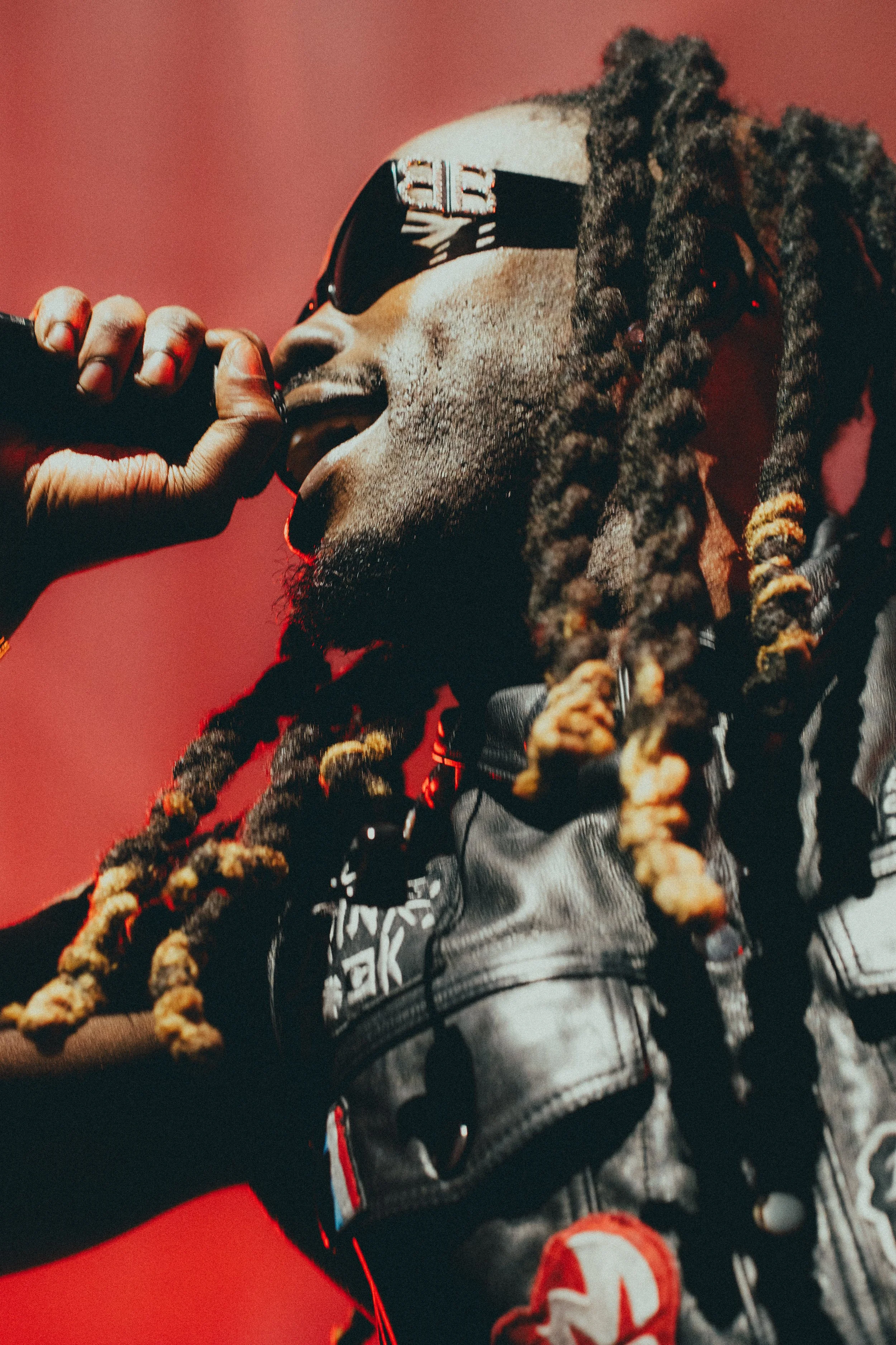 Un homme avec des dreadlocks porte des lunettes de soleil, tient un micro près de la bouche et porte une combinaison ou un vêtement de style urbain avec des motifs et des patchs, contre un fond rouge.