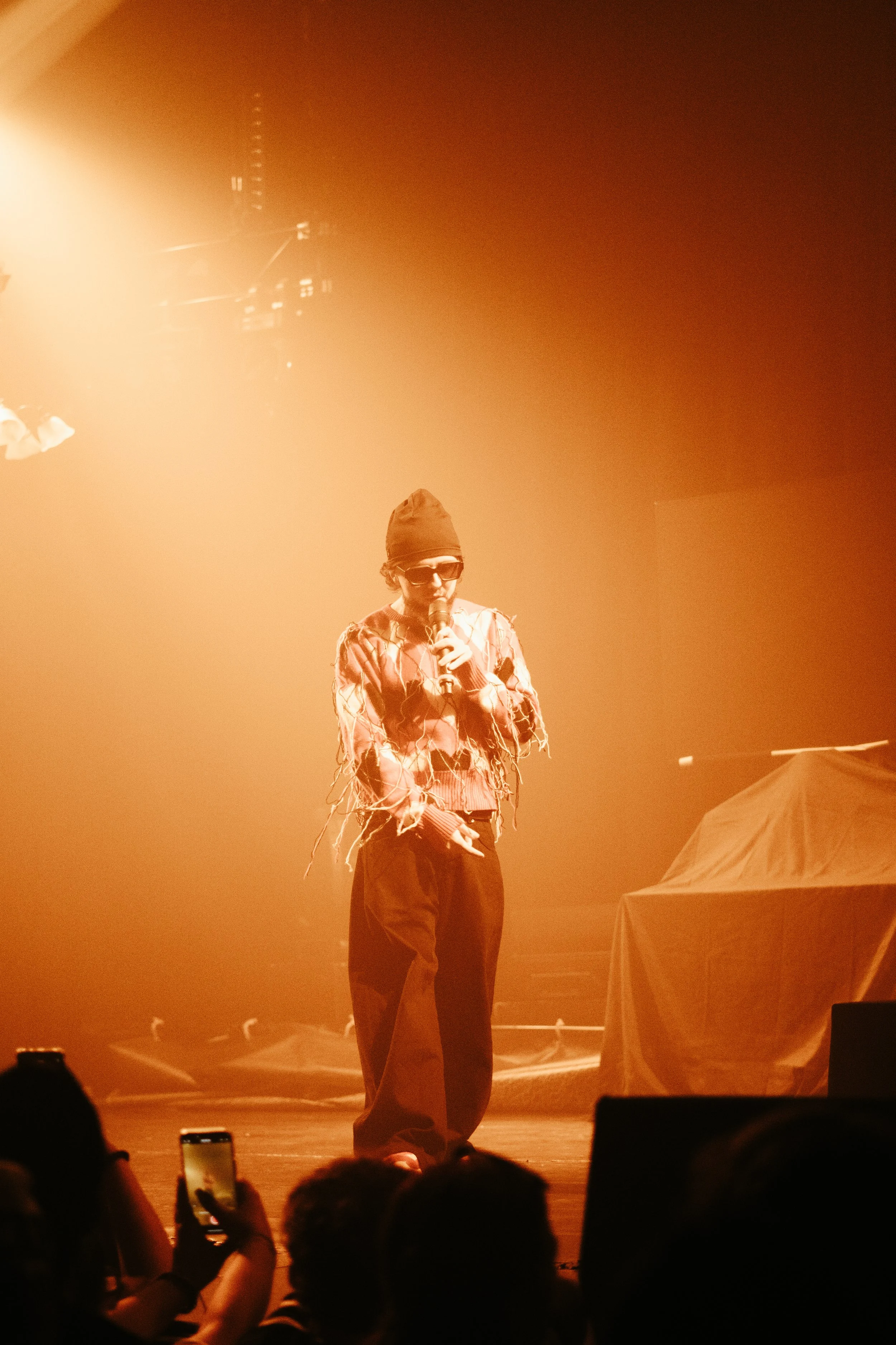 Une personne portant une casquette, des lunettes de soleil et un vêtement coloré chante ou parle dans un microphone sur scène, avec une lumière orange intense et un public en avant-plan.