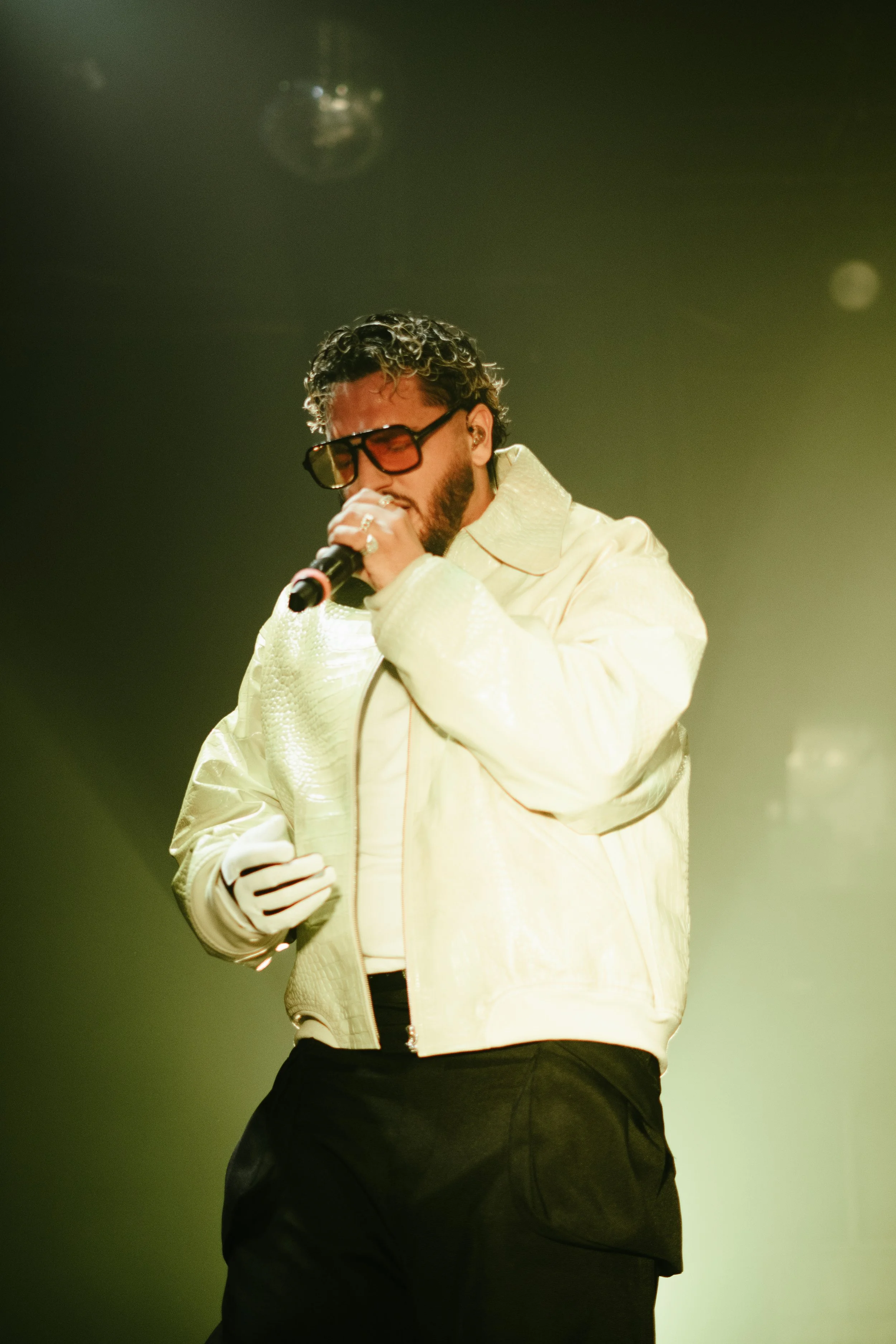 Un homme vêtu d'une veste beige claire, portant des lunettes de soleil, chante dans un microphone, sur scène avec un éclairage tamisé en arrière-plan.