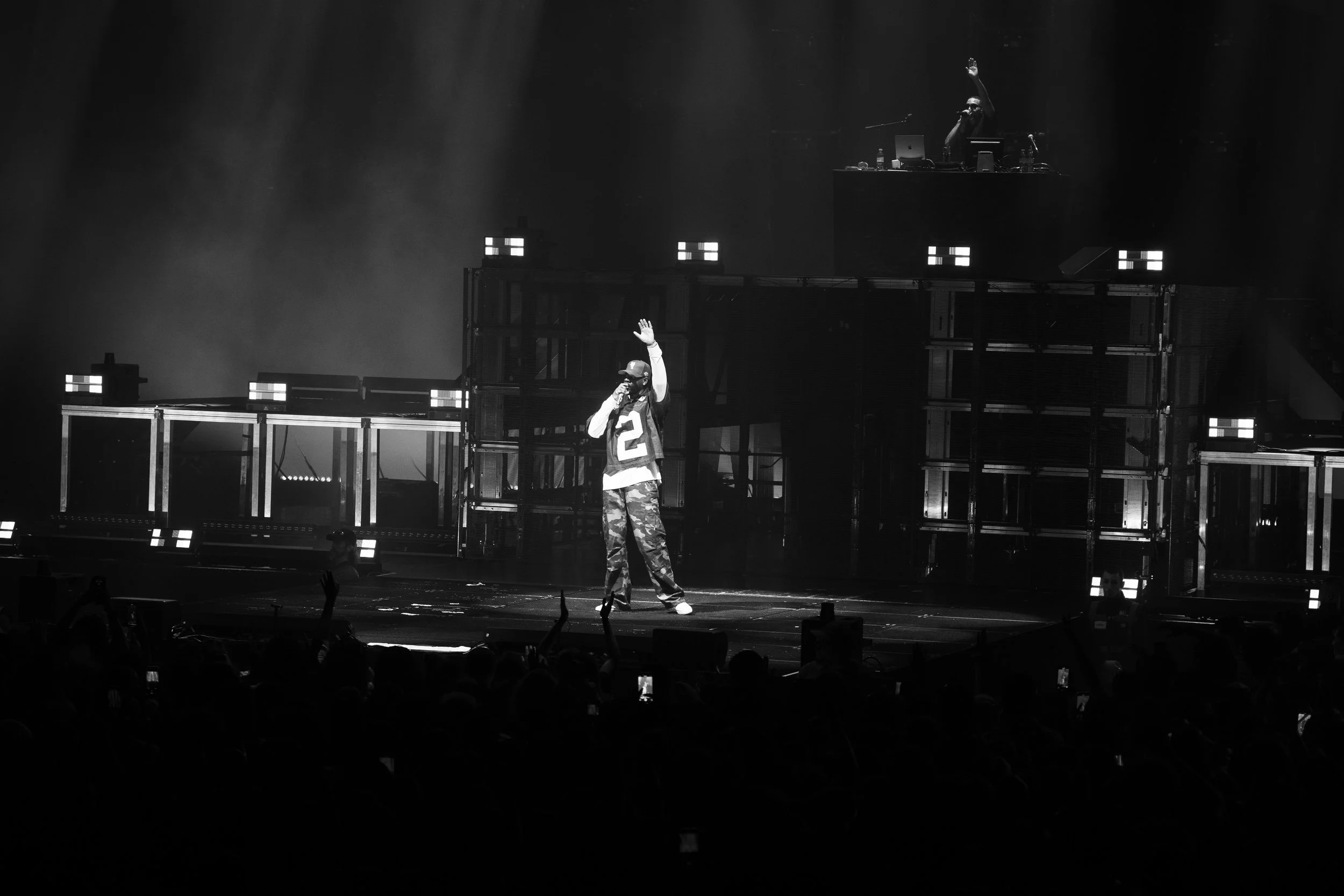 Interprète sur scène lors d'un concert, portant un maillot fluide avec le numéro 2, en tenue décontractée, avec un DJ derrière lui sur une plateforme et un public en bas, dans une ambiance sombre et lumineuse.