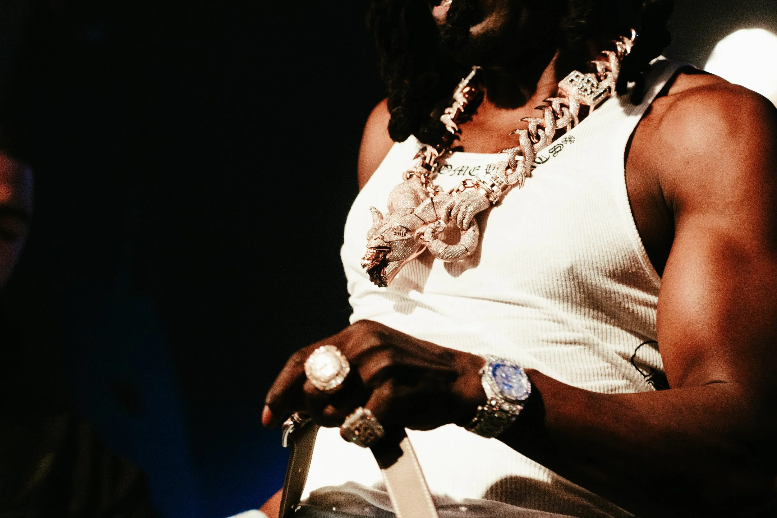 Une personne portant plusieurs bijoux en or, notamment une chaîne avec un pendentif en forme de dragon, un bracelet, une montre, et plusieurs bagues, portant un débardeur blanc.