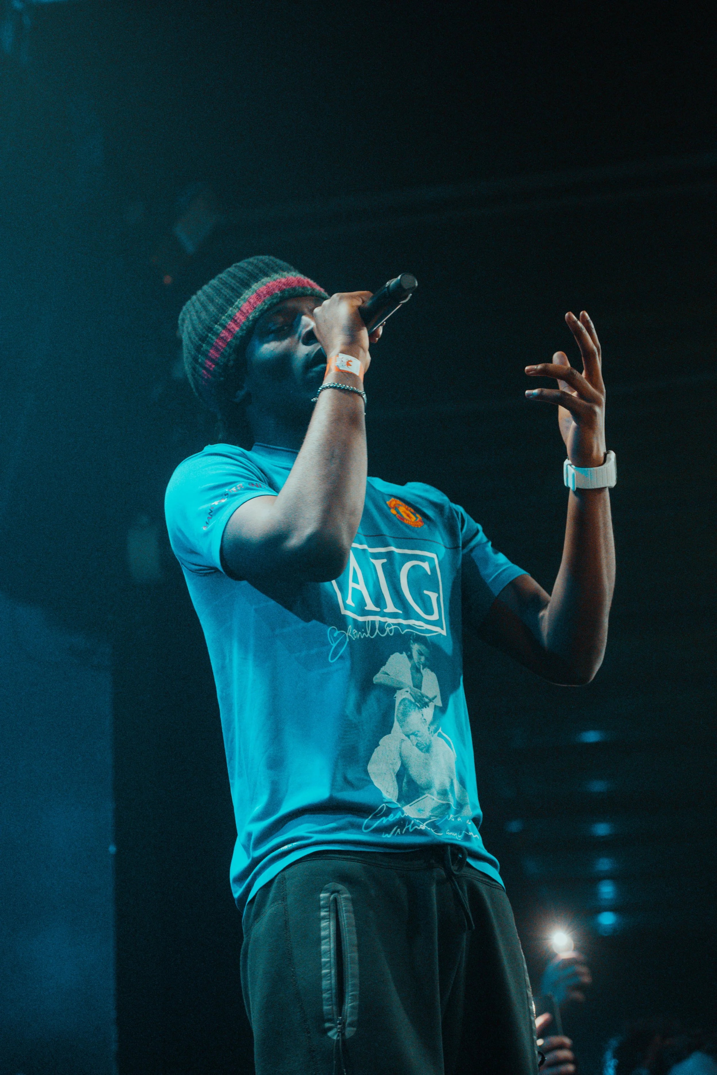 Une personne chante en utilisant un microphone sur scène, portant un bonnet gris à rayures rouges et une chemise bleue avec un logo de Manchester United.