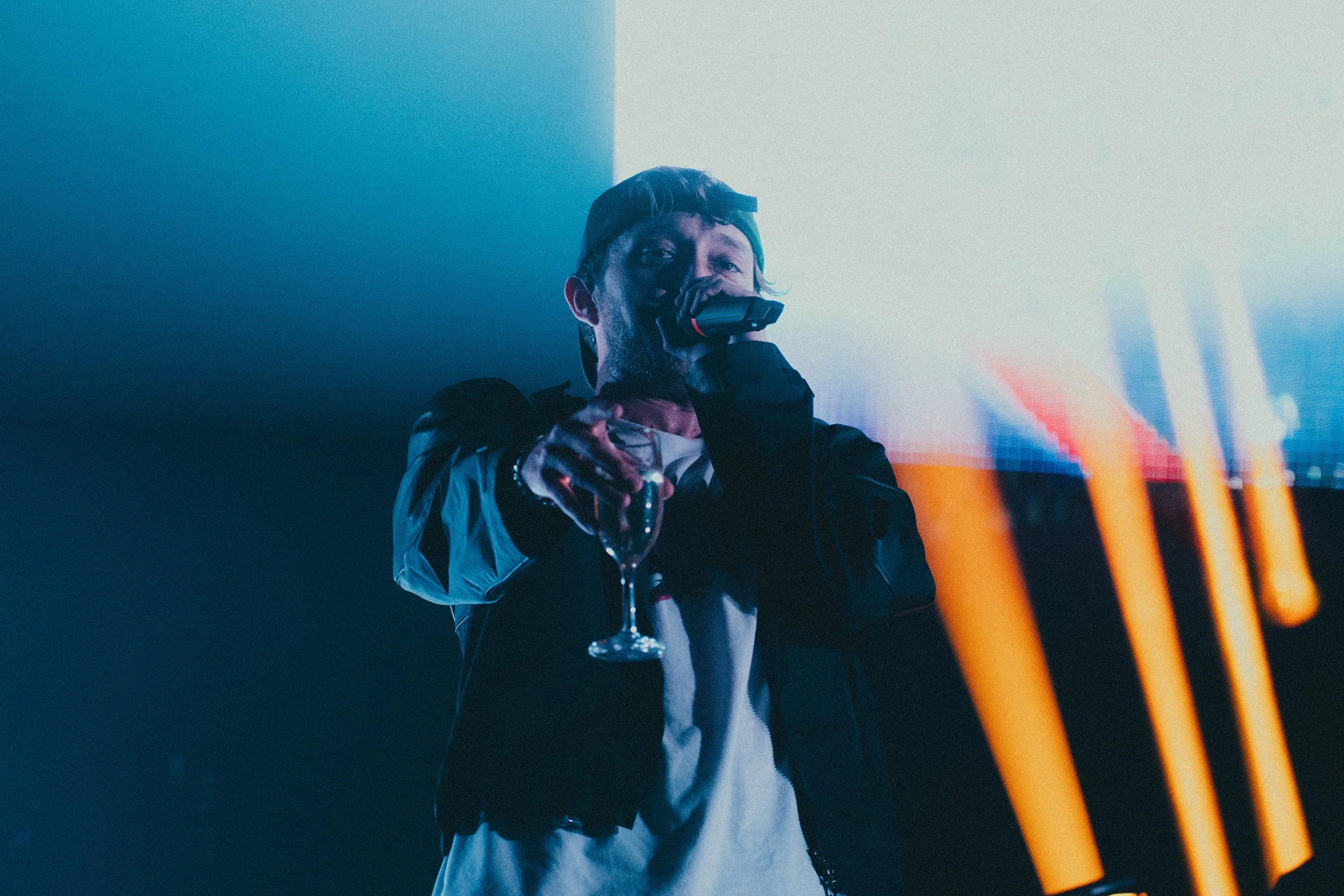 Un homme portant une casquette, un blouson noir, et des gants, tenant un verre de champagne et un micro, lors d'une fête ou d'un événement dans une ambiance sombre avec des lumières bleuâtres et orange.
