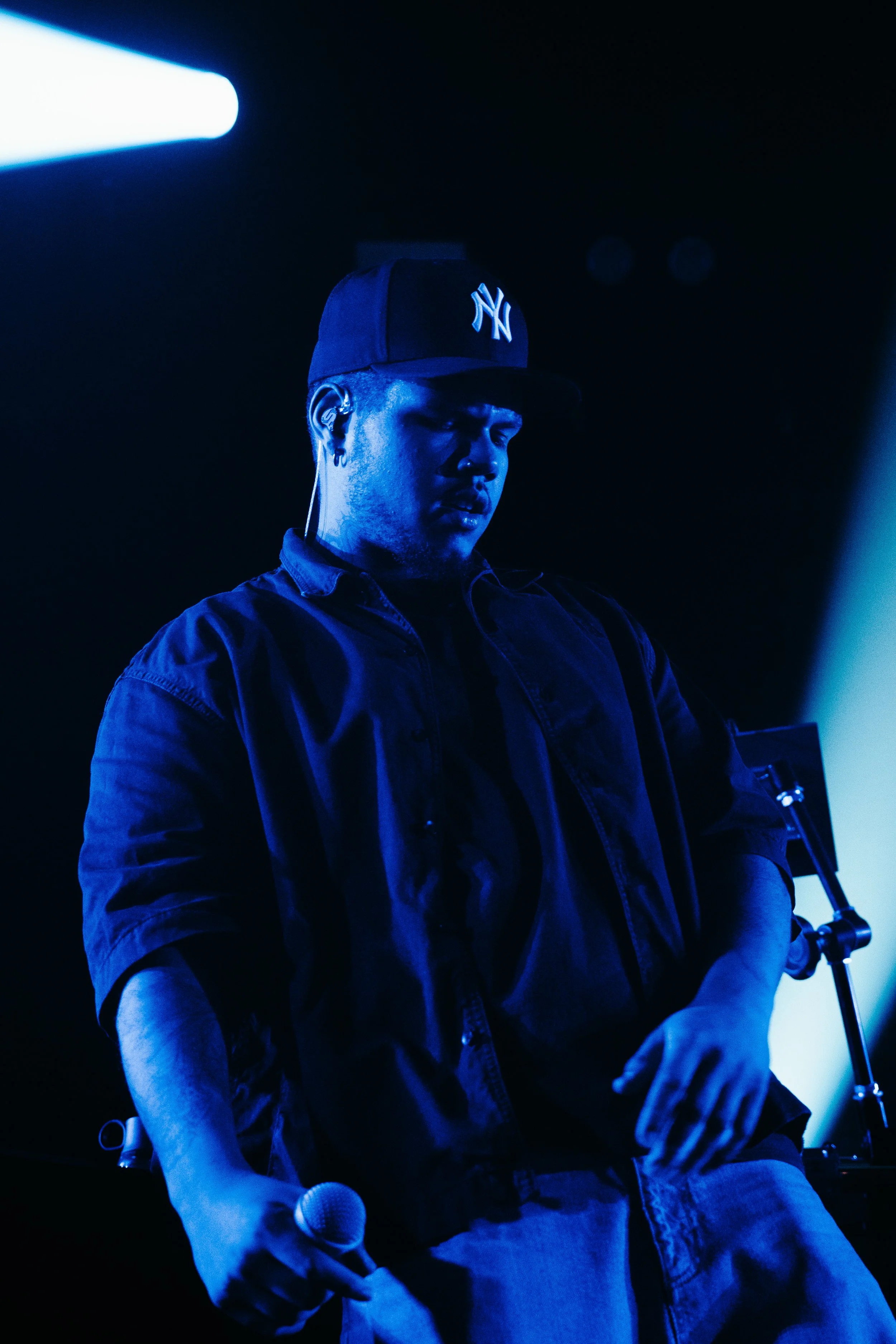 Un homme portant une casquette New York Yankees, tenant un micro, sous un éclairage bleu lors d'une performance musicale.