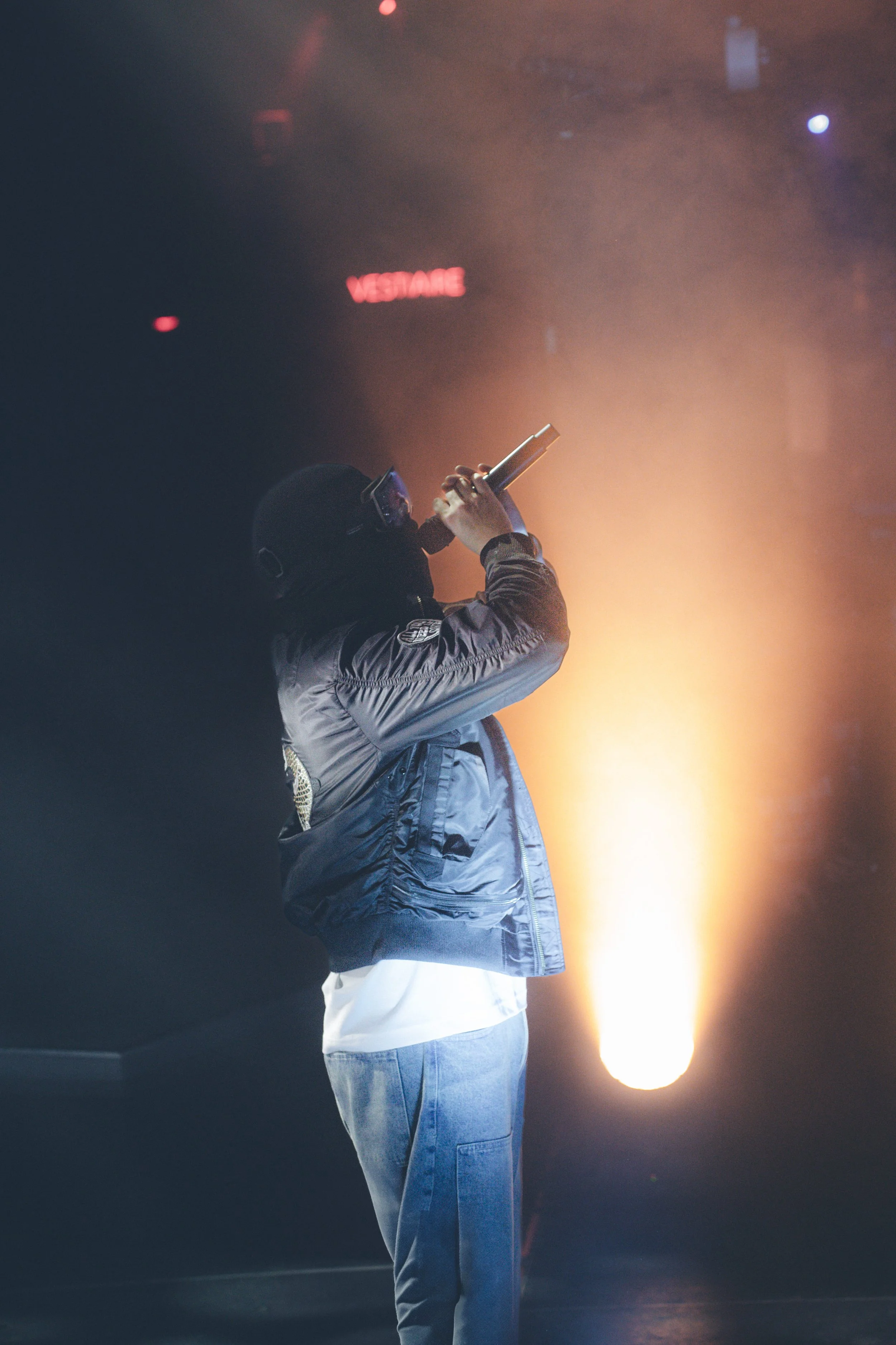 Une personne portant un casque, une veste noire et un jean, tenant un objet qui ressemble à un pistolet, sous un éclairage chaud, dans un environnement sombre avec une lumière rouge en arrière-plan.