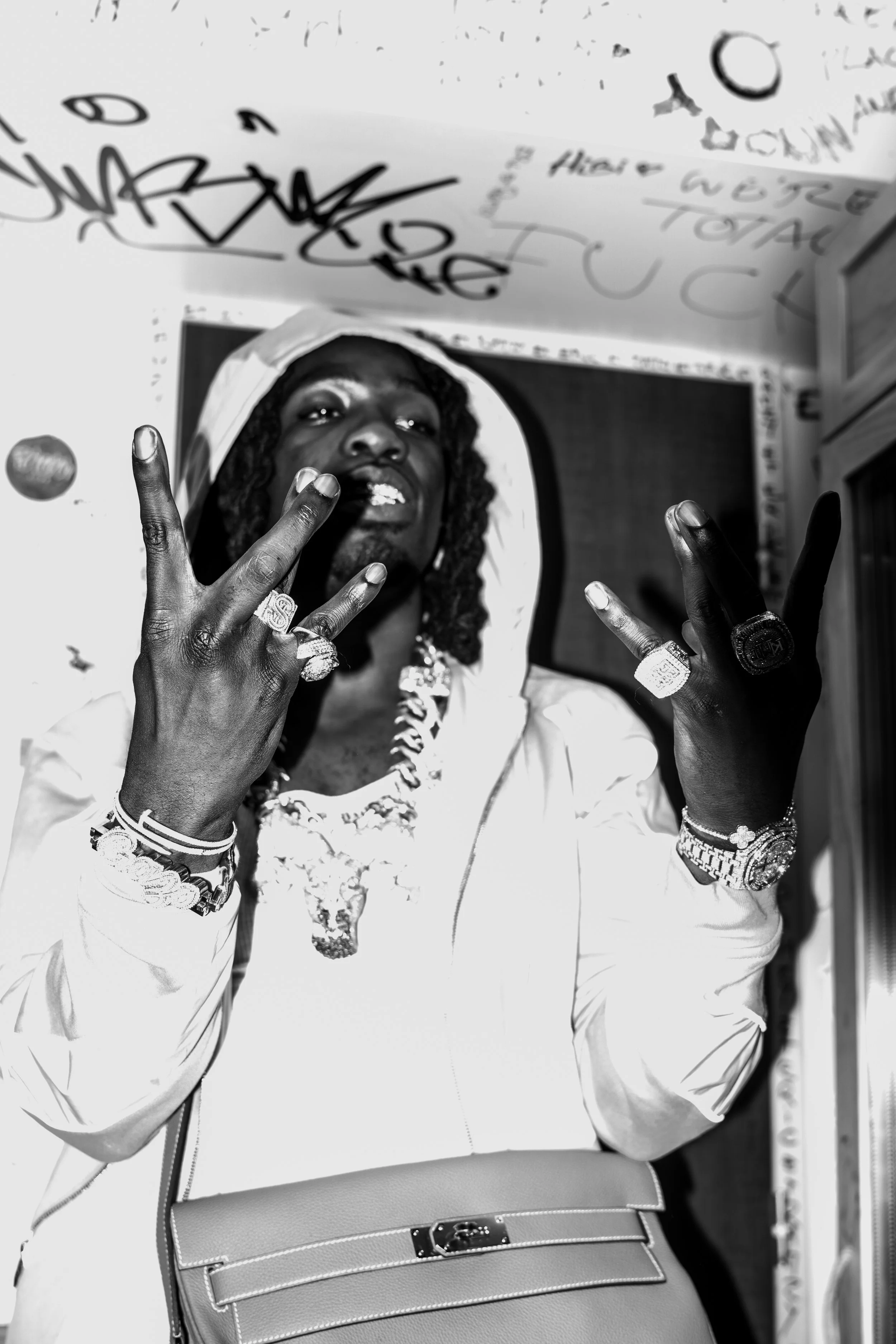 Photo en noir et blanc d'un homme portant des bijoux et des vêtements de style bling-bling, faisant un signe de la main devant un mur avec des graffitis.