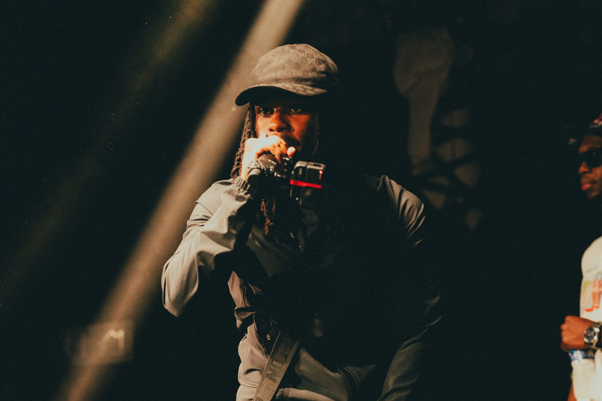 Une personne portant une casquette noire sombre et veste noire, tenant un microphone et un petit appareil de prise de vue, dans une scène sombre avec un éclairage focalisé.