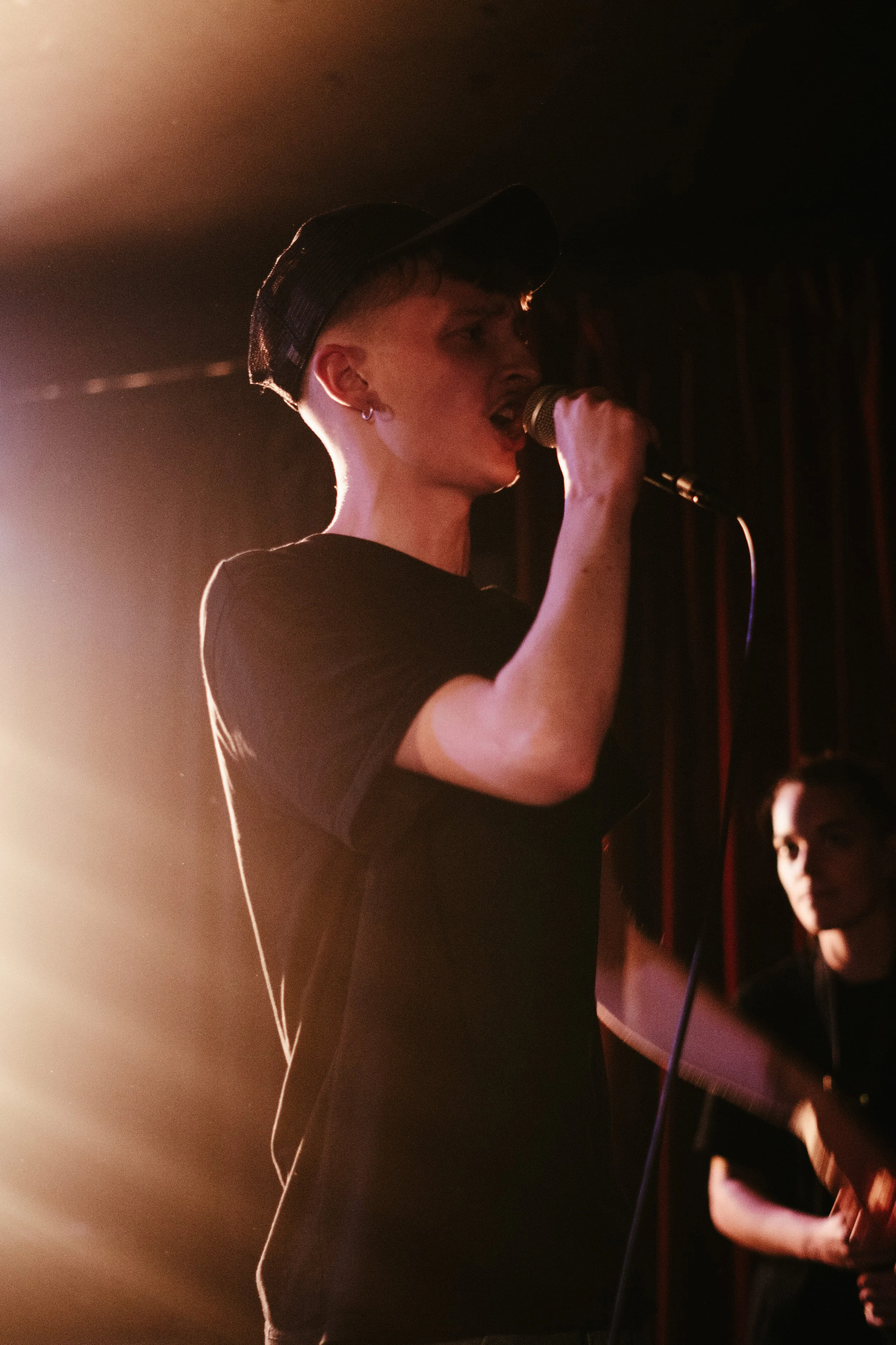 Jeune chanteur portant une casquette, chantant dans un micro lors d'une performance sur scène avec un musicien en arrière-plan.