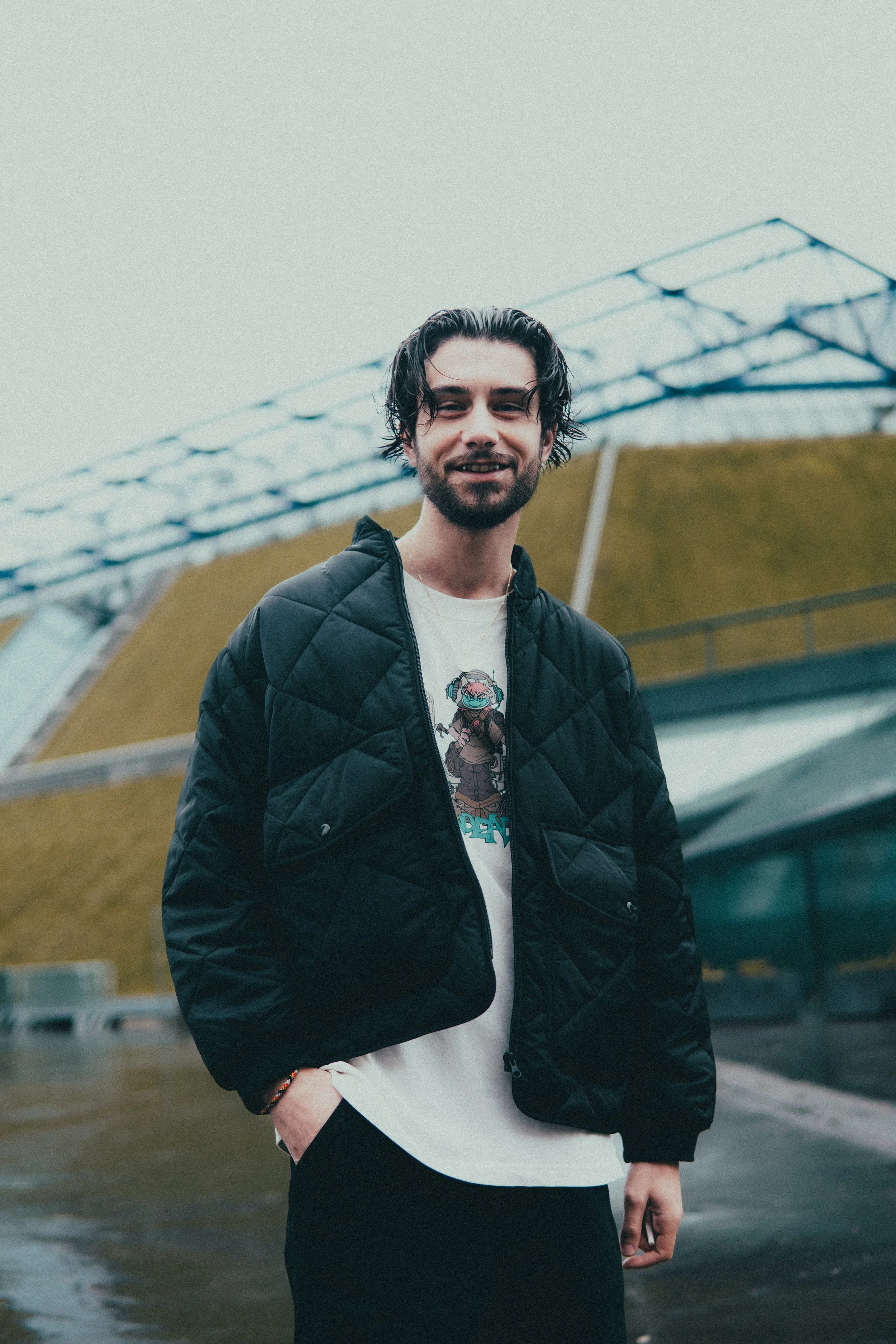 Un jeune homme souriant posant en extérieur, portant une veste noire et un t-shirt avec un dessin animé, avec un bâtiment moderne en arrière-plan.
