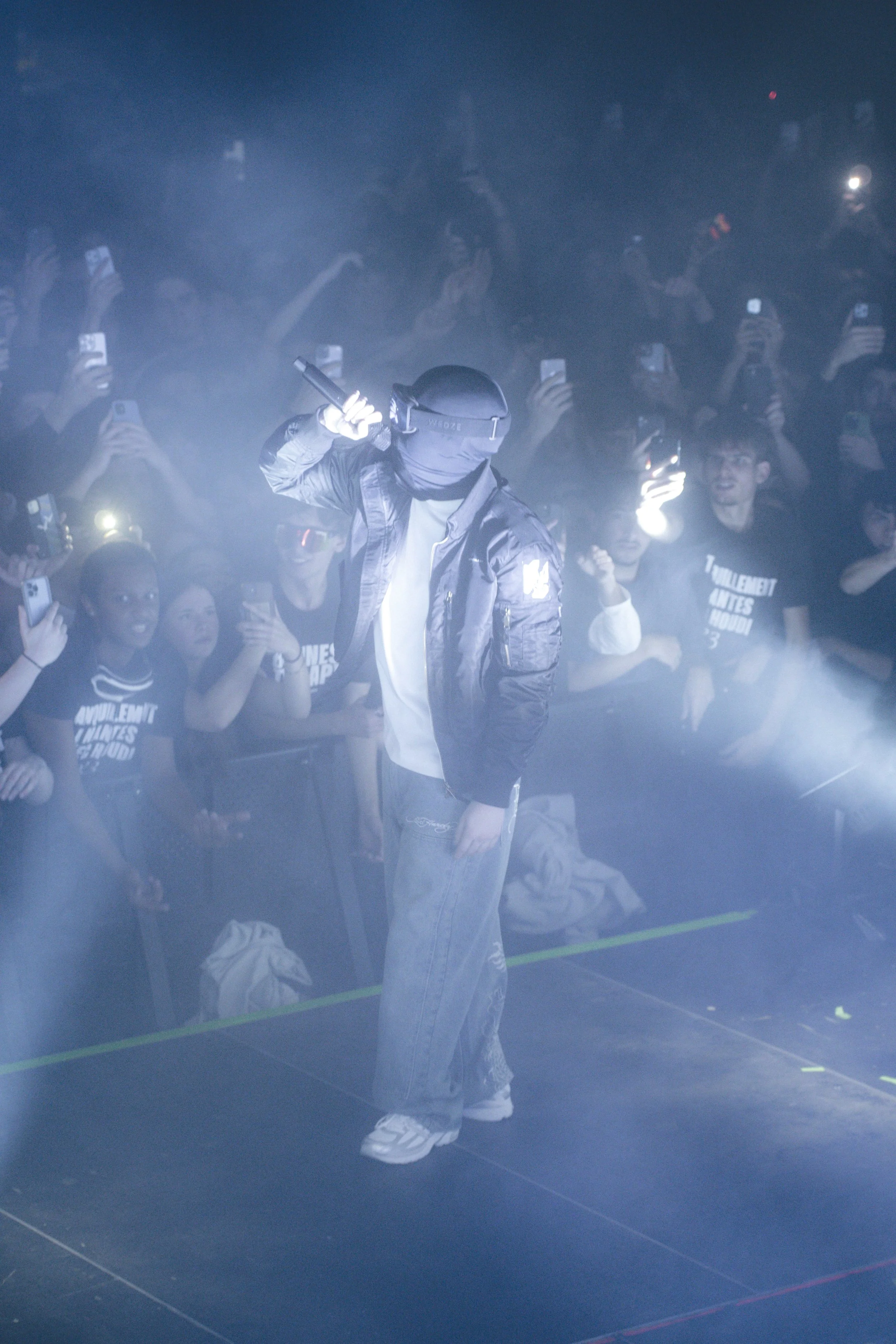 Un artiste de rap sur scène en tenue décontractée, avec une veste en cuir et un casque, dans une ambiance brumeuse. La foule l'encourage et prend des photos avec leurs téléphones.