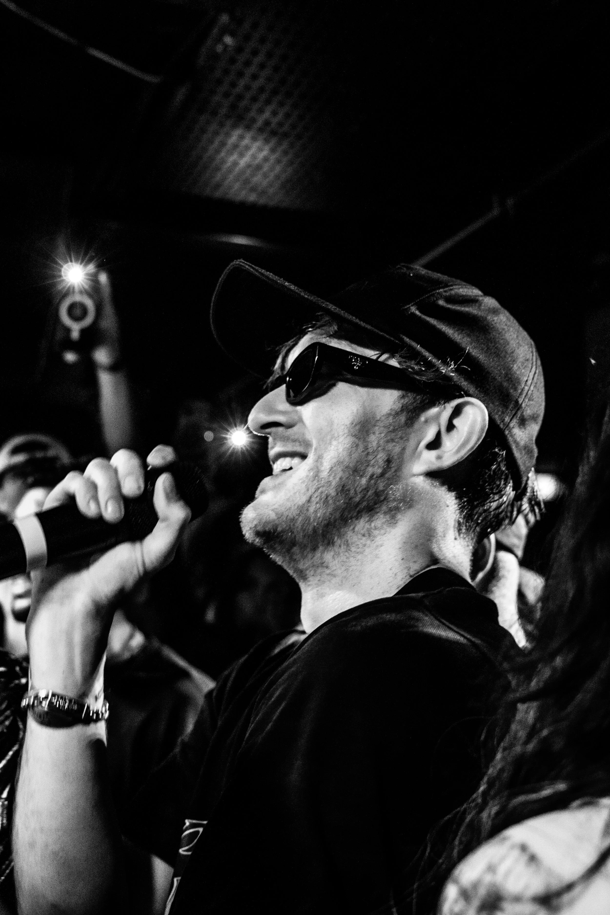 Homme souriant portant des lunettes de soleil, un casquette et tenant un microphone, dans une ambiance sombre avec des lumières.