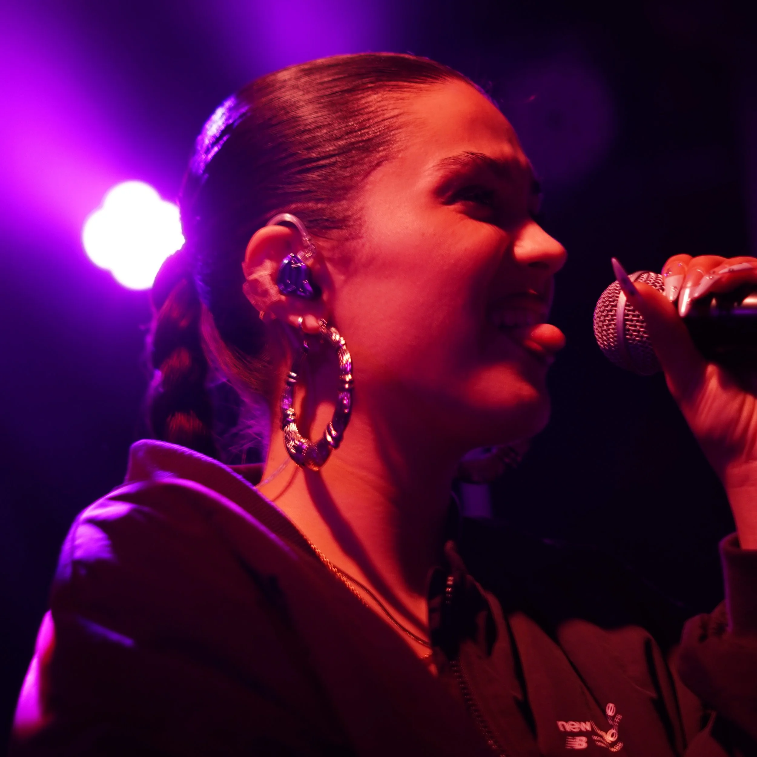 Femme chantant ou parlant dans un microphone lors d'une performance, éclairage violet et rose en arrière-plan.