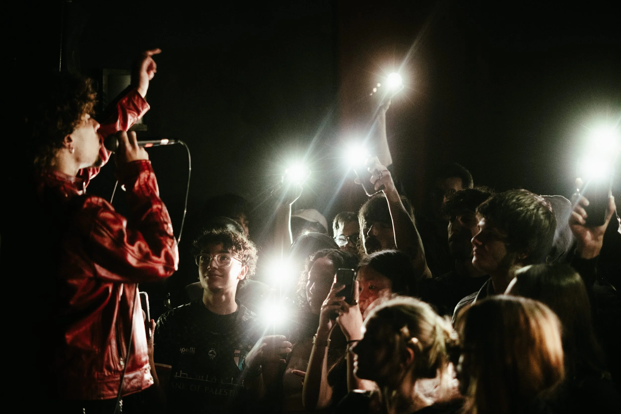 Une personne chante avec un microphone dans un environnement sombre, entourée de plusieurs autres personnes tenant des téléphones portables avec flash allumé, capturant l'instant.