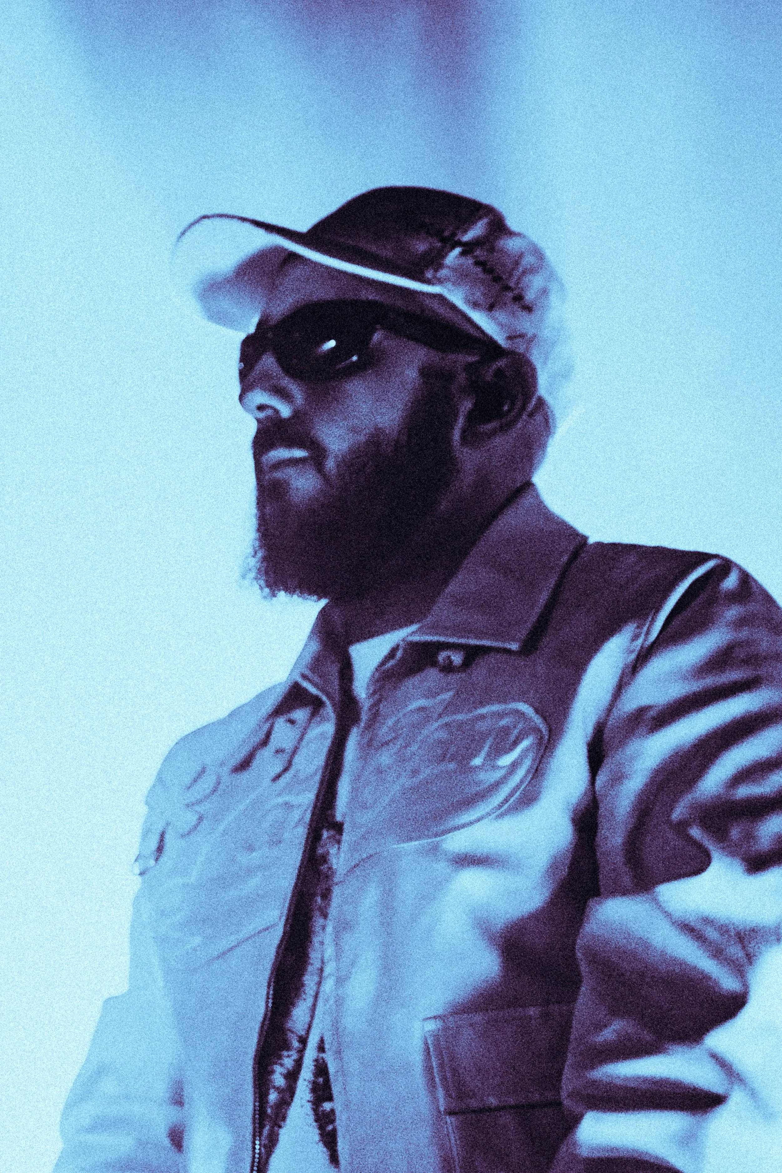 Un homme portant des lunettes de soleil, un chapeau et une veste en cuir, regardant vers la gauche sous un éclairage bleu