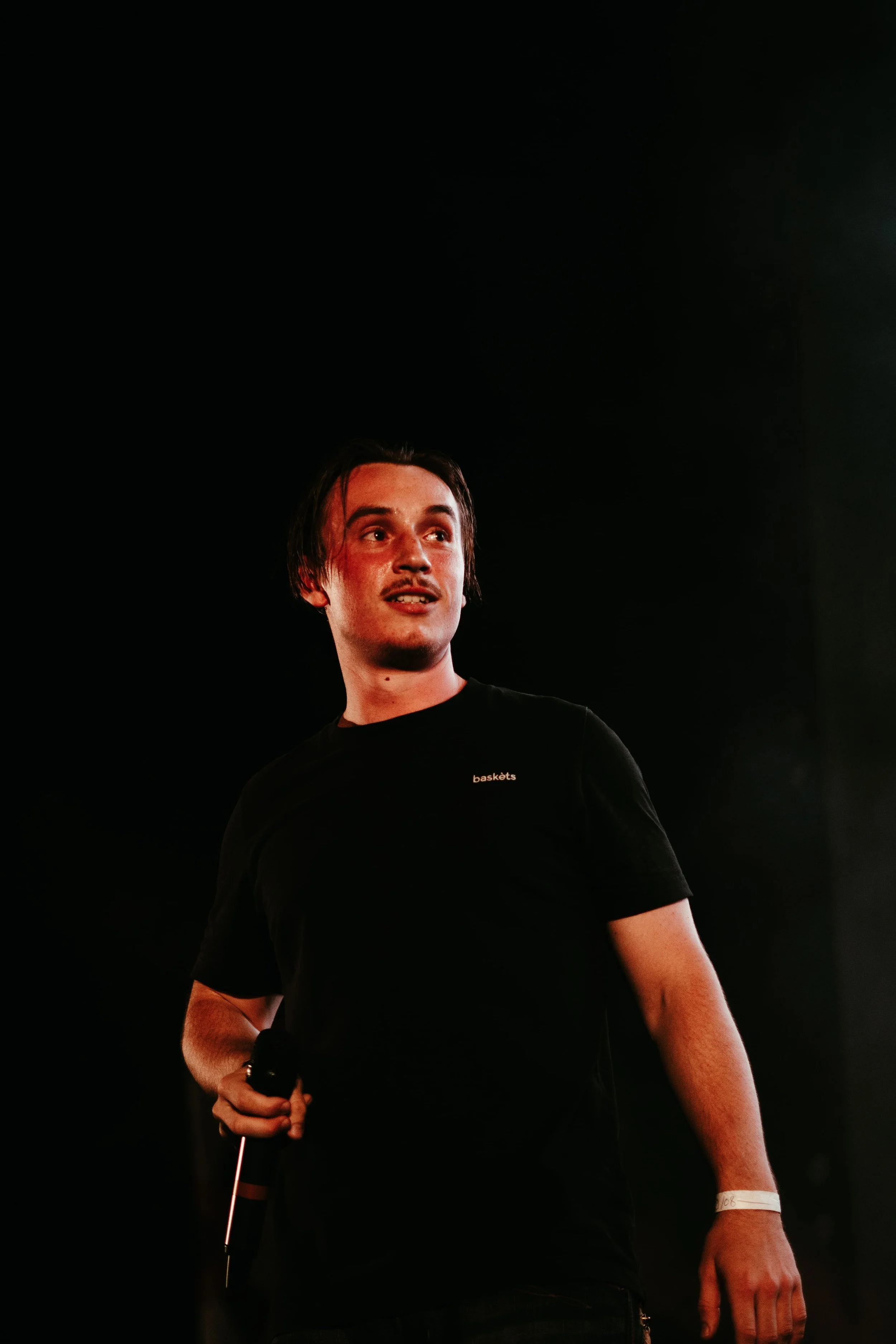 Un jeune homme tient un microphone, portant un t-shirt noir avec le mot 'baskets' dessus, sur scène, avec un fond sombre.