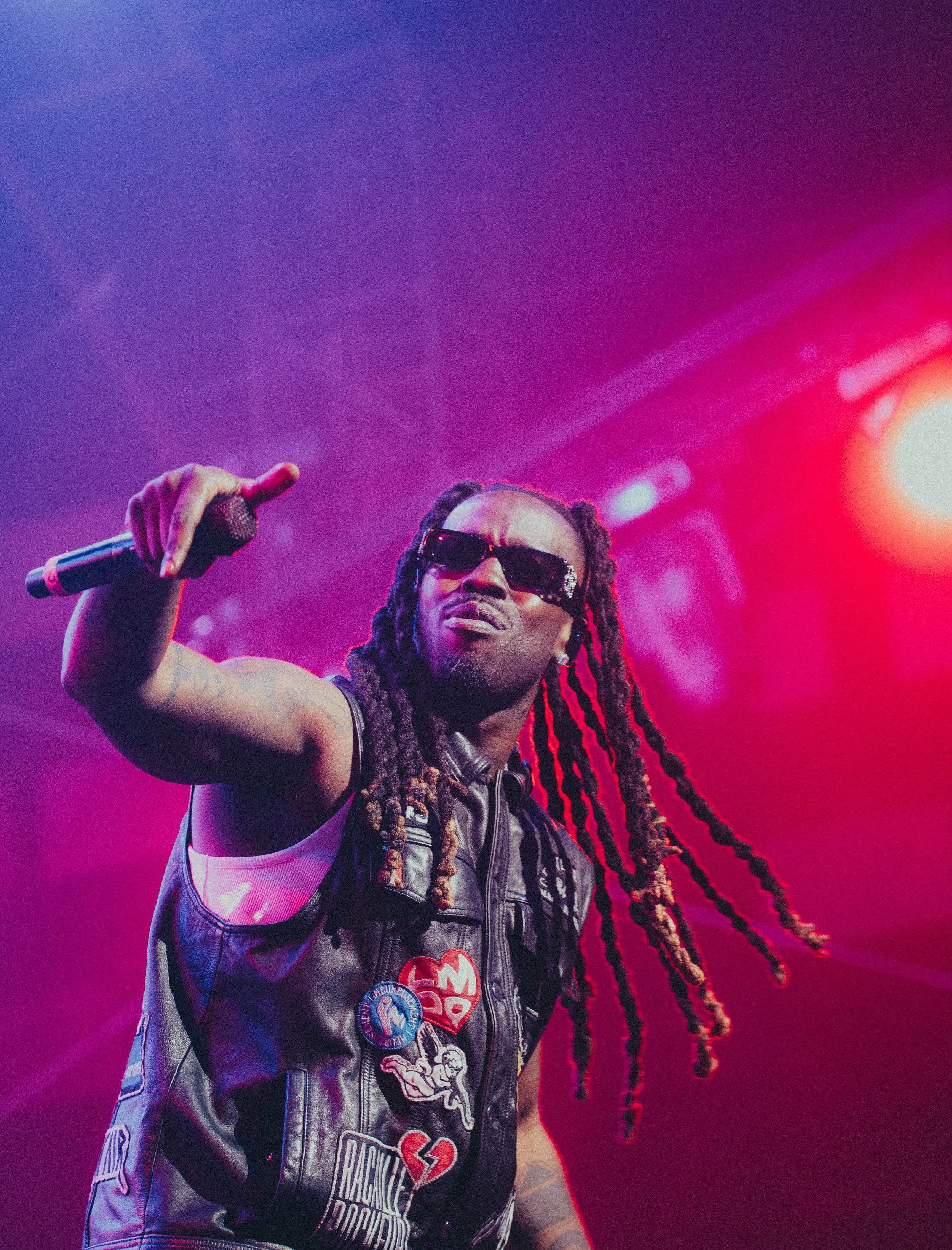 Un chanteur avec de longs dreadlocks portant des lunettes de soleil et une veste en cuir, tenant un microphone sur scène avec des lumières colorées en arrière-plan.