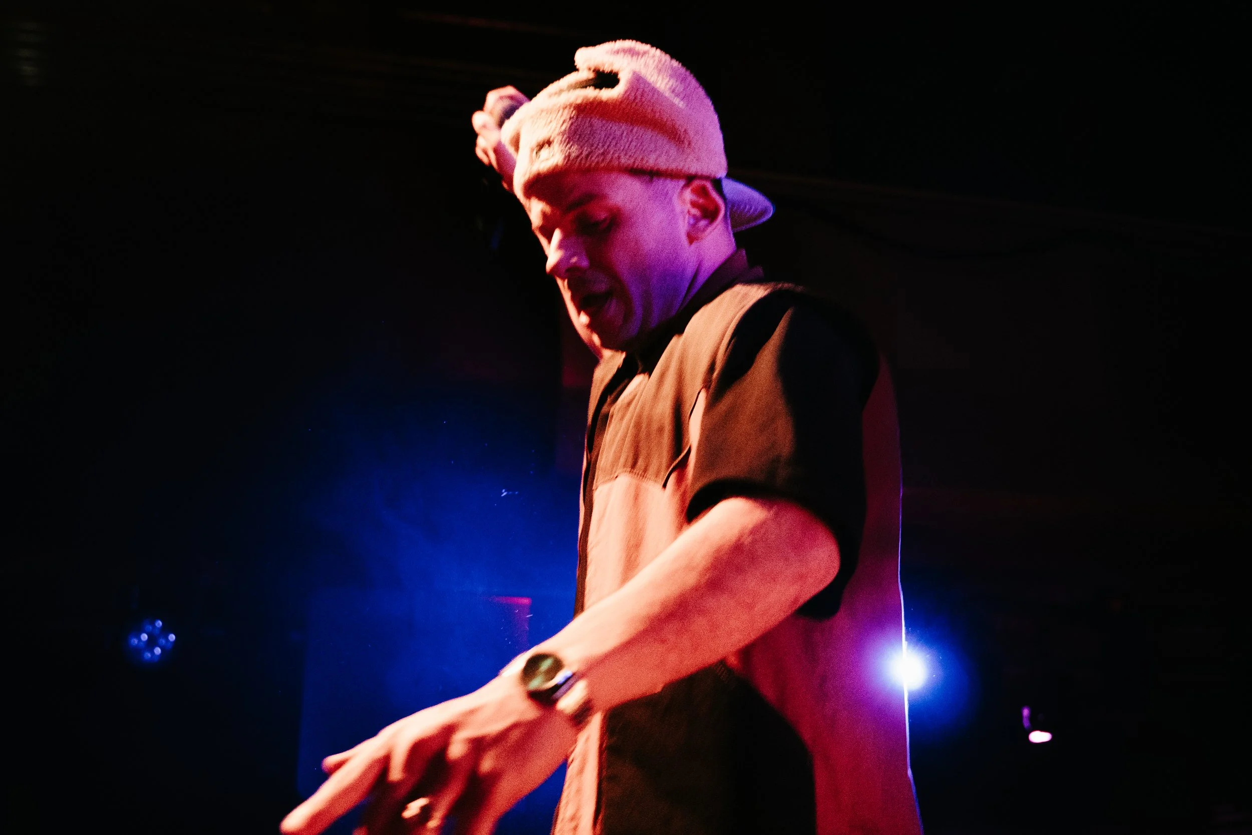 Un homme portant un bonnet beige, une chemise à manches courtes et une montre, en train de jouer de la musique sur scène avec un arrière-plan sombre et des lumières colorées.