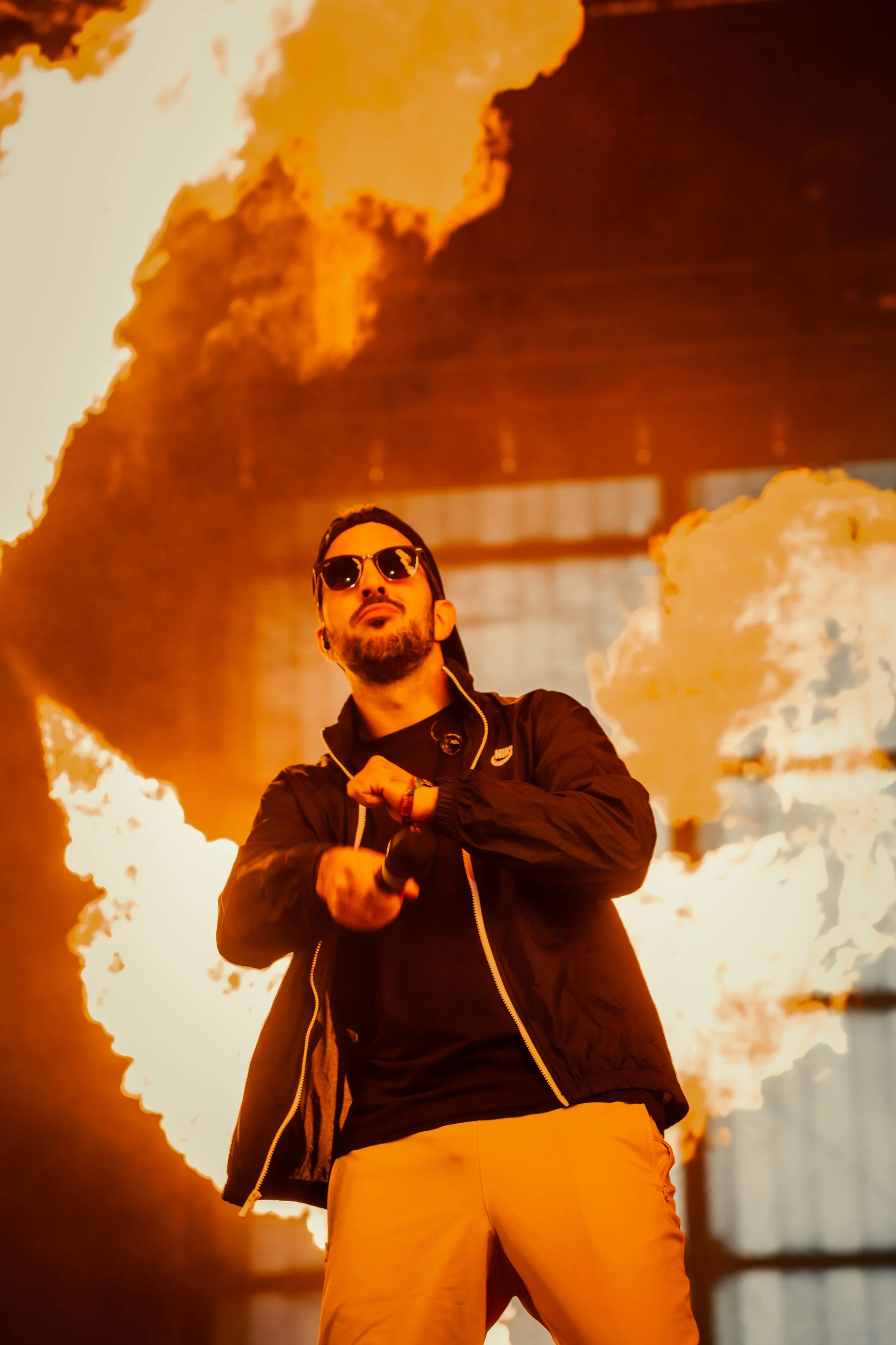 Un homme portant des lunettes de soleil, une veste noire et un t-shirt noir, tenant un microphone, avec un feu de grande ampleur derrière lui.
