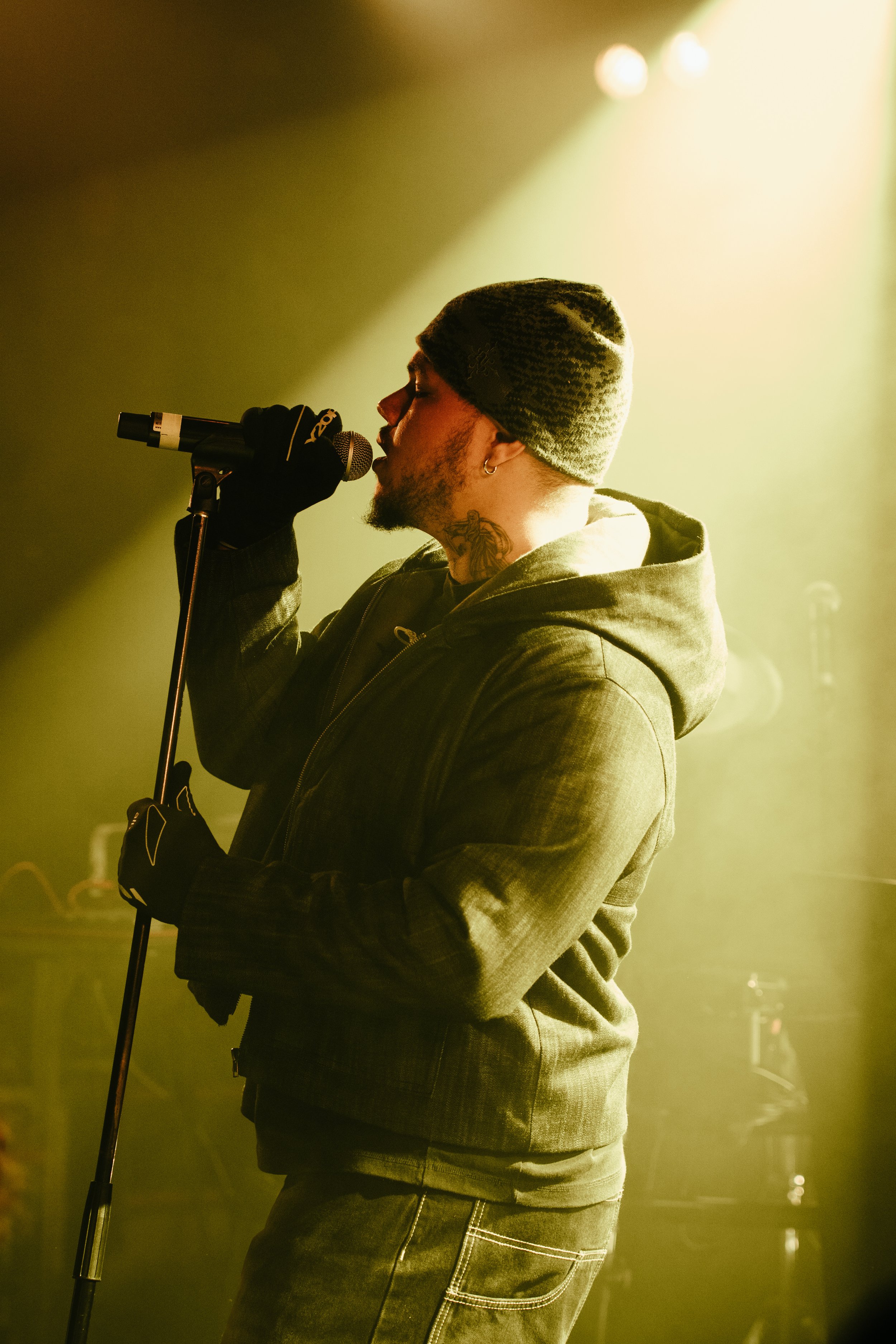 Un homme chante dans un microphone, portant un bonnet et un hoodie, sous un éclairage jaune.
