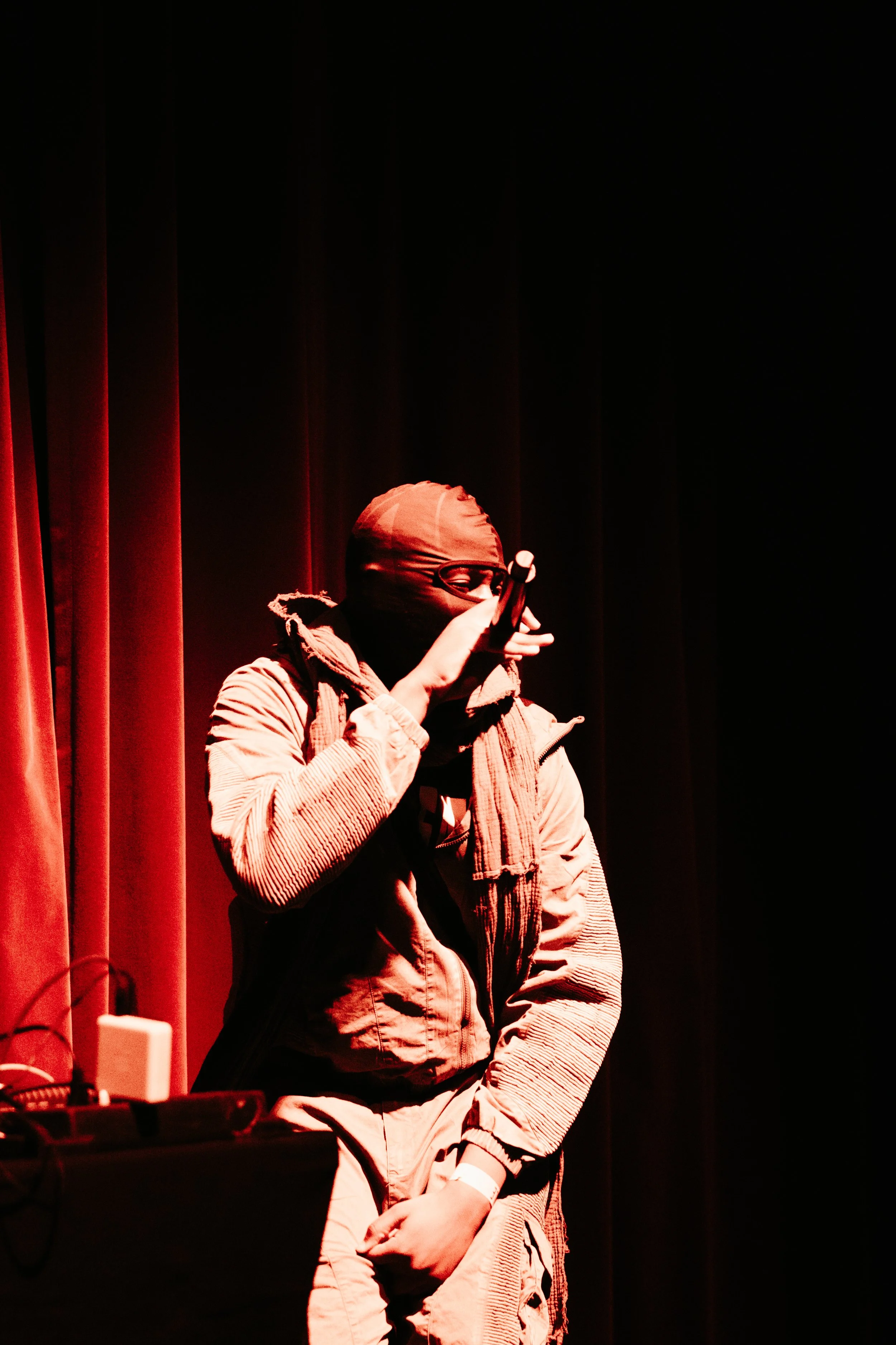 Un artiste de rue portant un masque, un bandeau sur la tête, et un blouson, se tenant devant un rideau rouge, utilisant un microphone dans un cadre sombre.