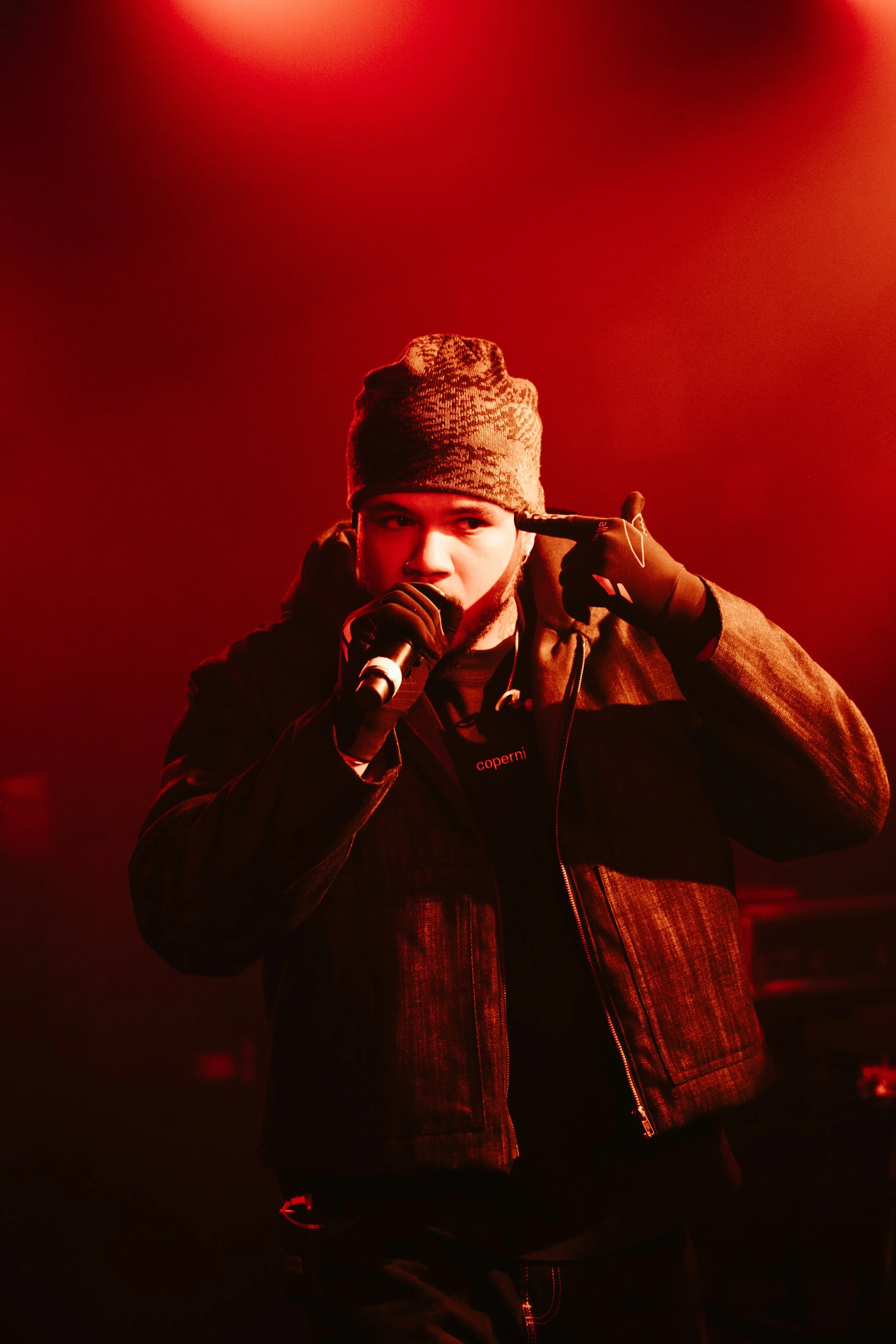 Jeune homme portant un bonnet gris, une veste noire, tenant un microphone, faisant un geste avec ses doigts près de la tête, éclairé par une lumière rouge.