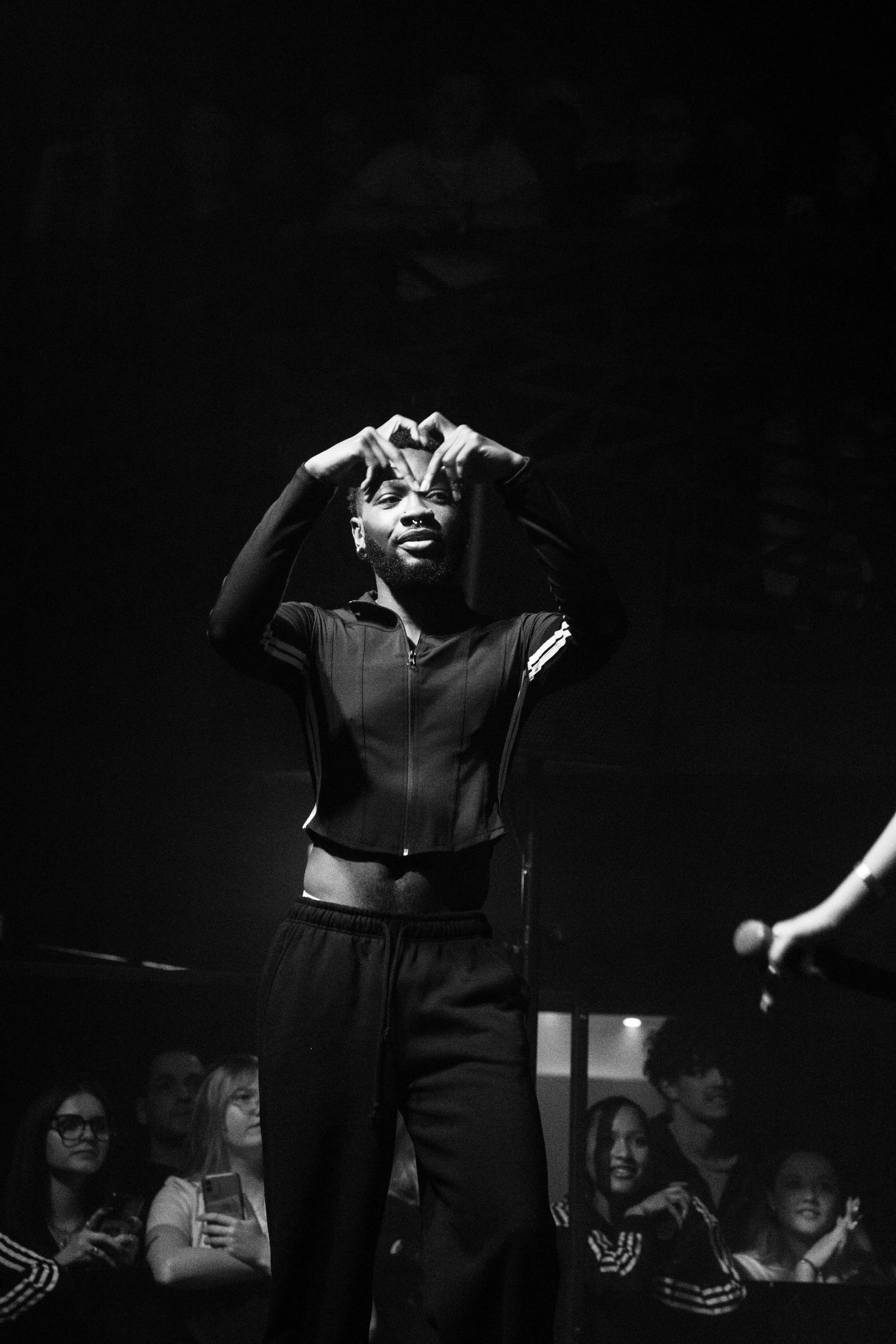 Une femme avec un crop top noir et un sweat à capuche fait un geste en forme de cœur avec ses mains sur scène lors d'un concert, avec un groupe de spectateurs en arrière-plan.
