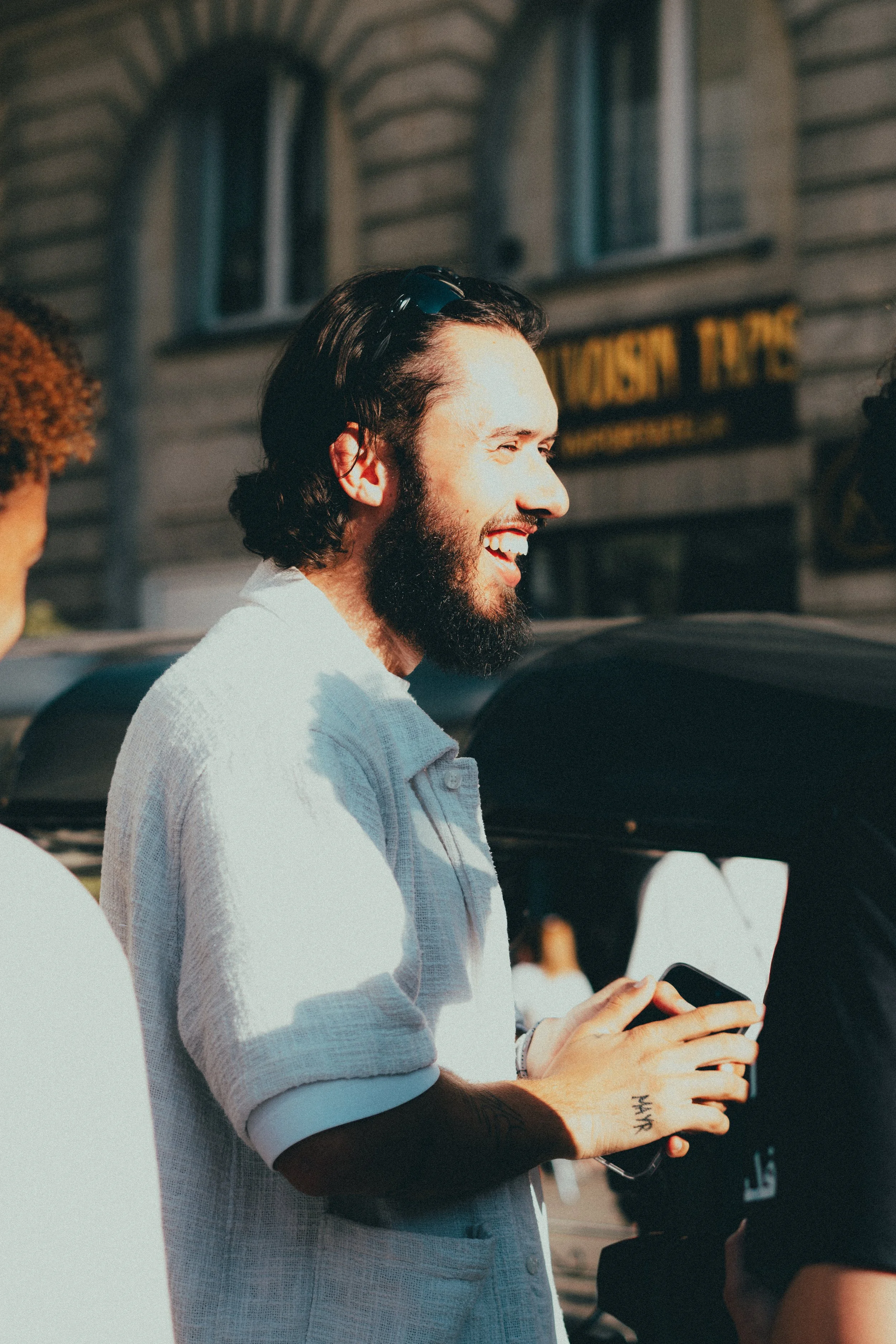 Jeune homme avec une barbe, souriant, tenant un téléphone, en plein air, ensoleillé, avec des bâtiments en arrière-plan.