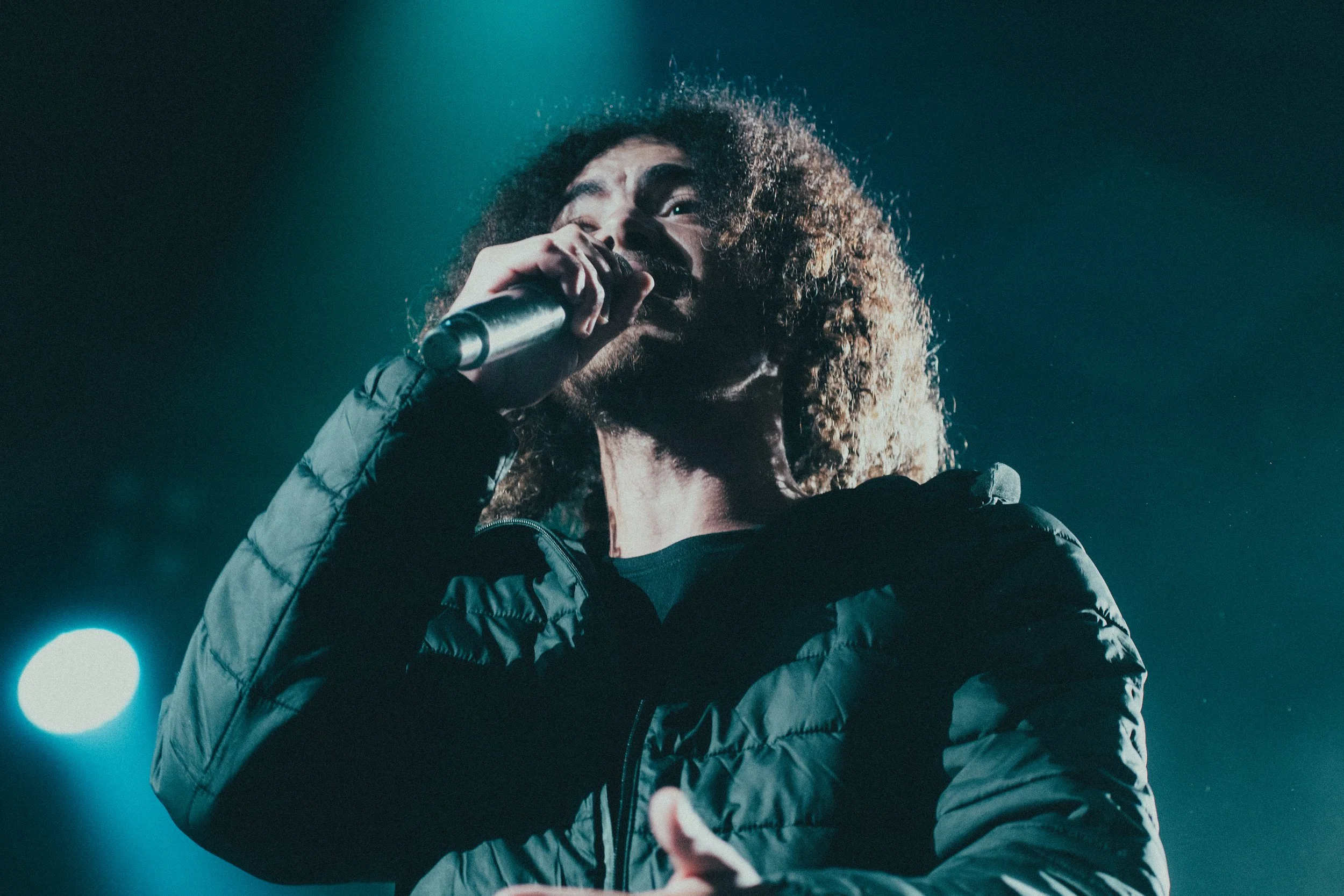 Un homme avec des cheveux bouclés et une barbe tient un microphone en chantant sur scène, éclairé par des lumières bleues.