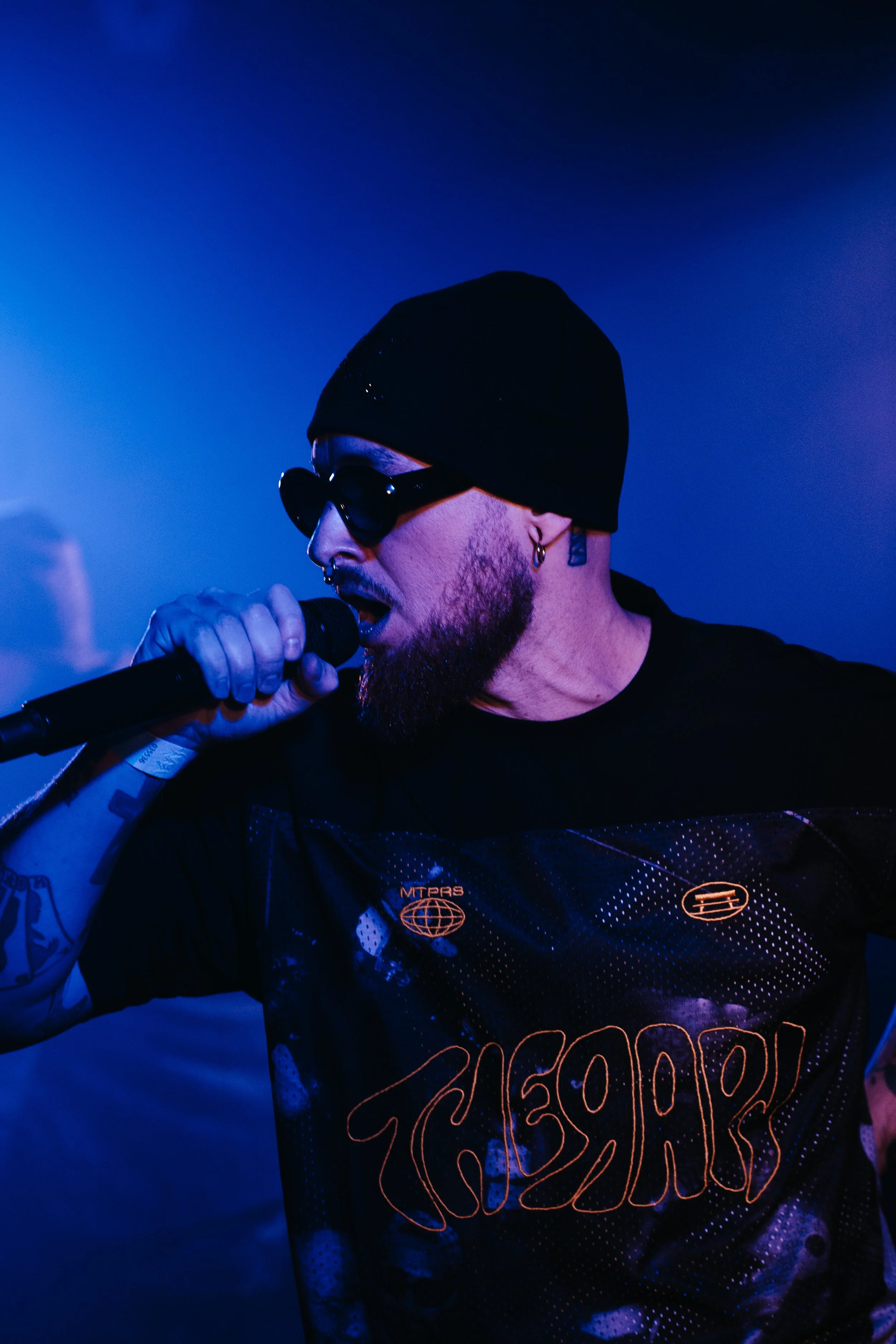 Un homme avec une barbe, portant un bonnet noir et des lunettes de soleil, chante dans un microphone dans un environnement sombre éclairé par une lumière bleue.