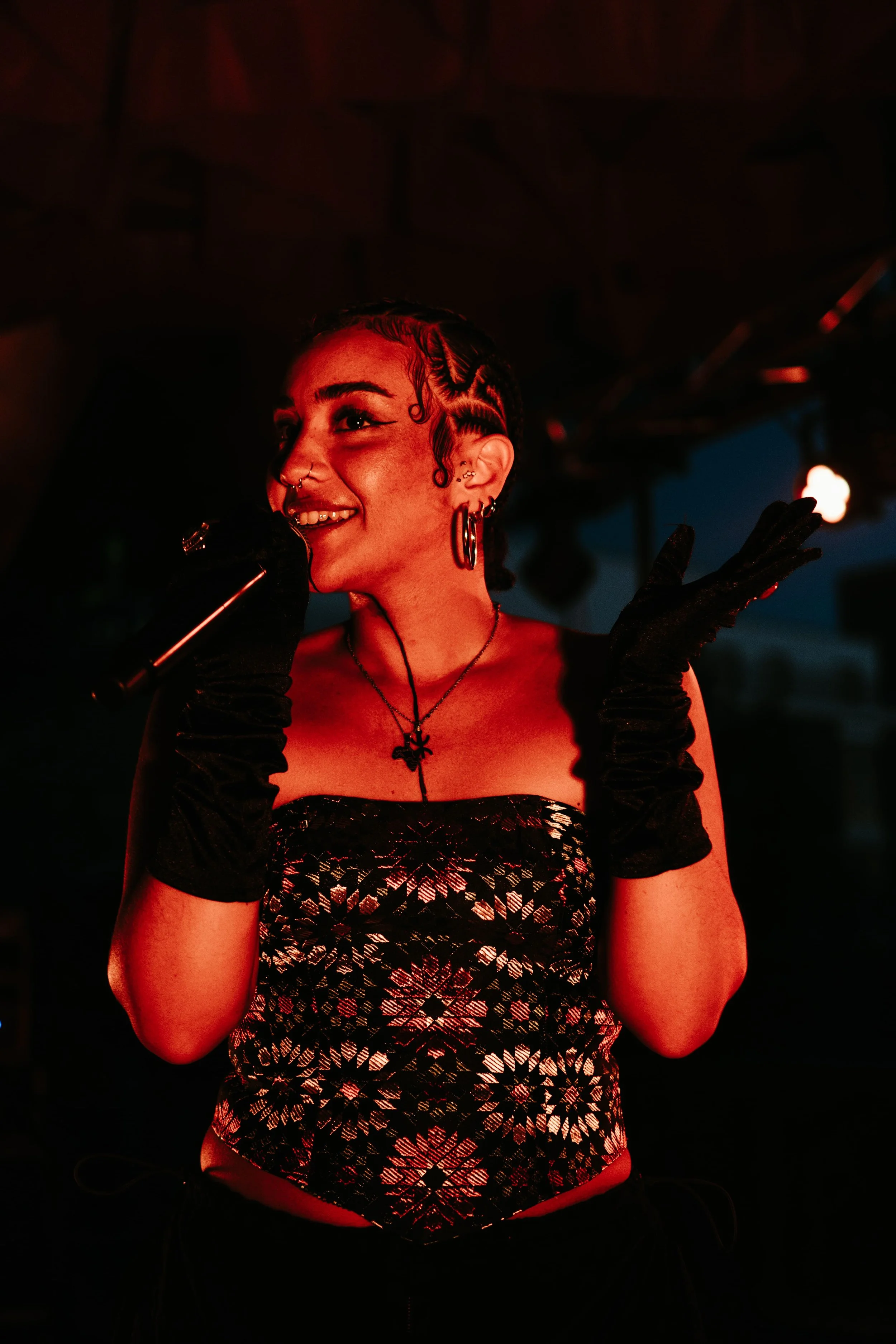 Une femme avec des cheveux bouclés, portant un corsage noir et rouge avec un motif en forme de fleurs, avec des boucles d'oreilles, un collier et un piercing au nez, tenant un microphone et souriant dans un environnement sombre avec un éclairage roug