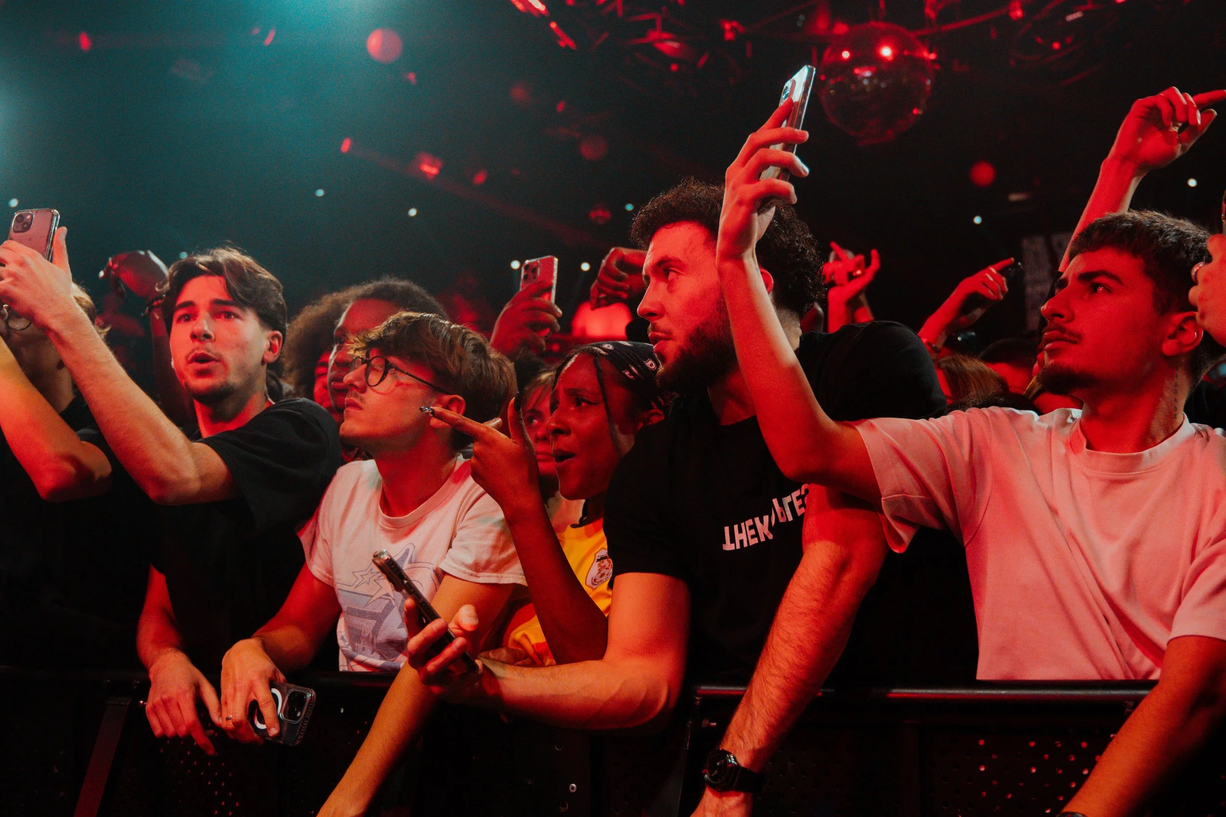 Groupe de jeunes spectateurs au premier rang lors d'un concert, certains prenant des photos ou vidéos avec leurs téléphones, sous un éclairage rouge et noir avec des ballons suspendus au plafond.