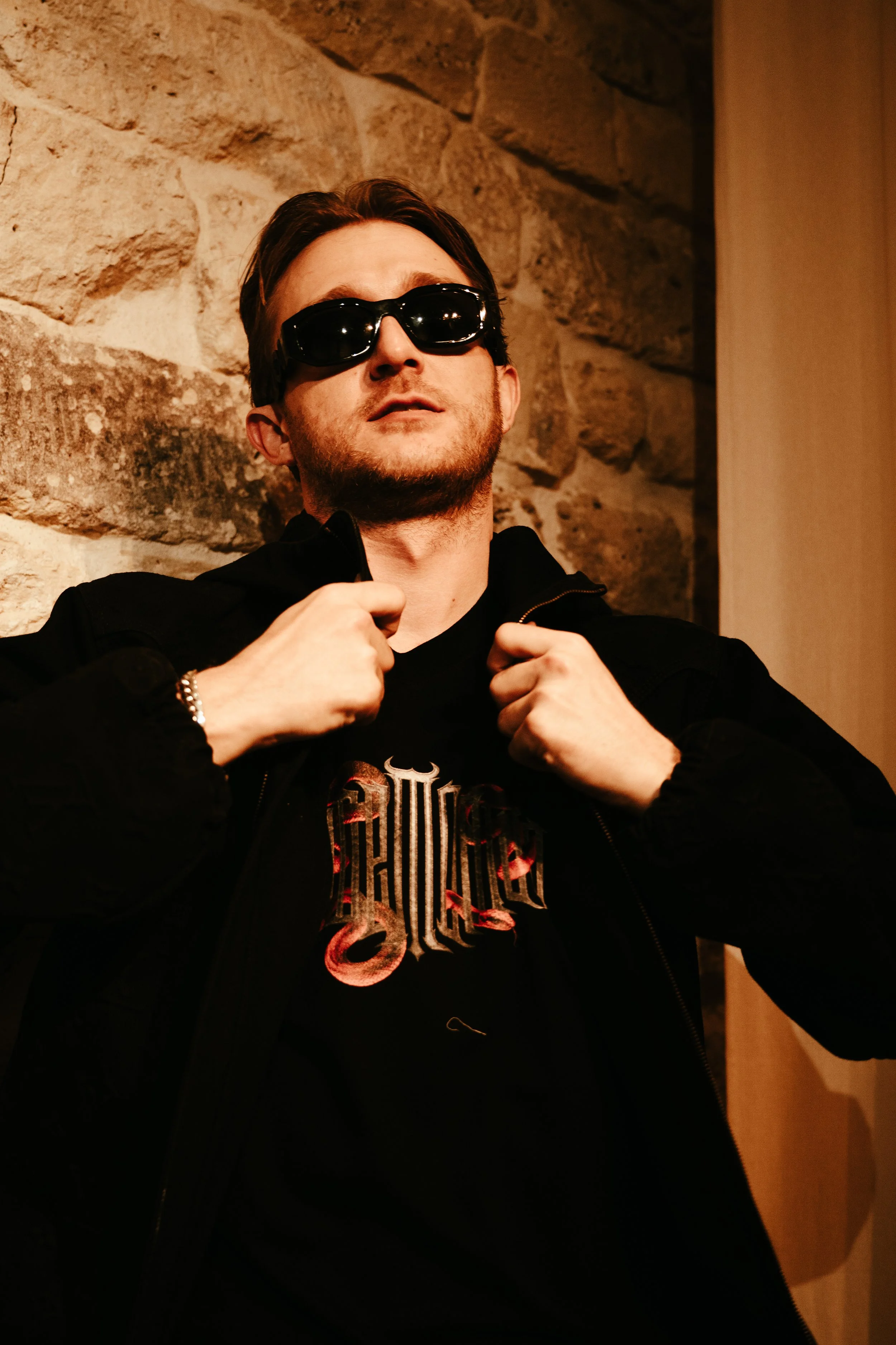Un homme portant des lunettes de soleil noires, une veste noire et un t-shirt avec un design graphique, pose devant un mur en pierre.