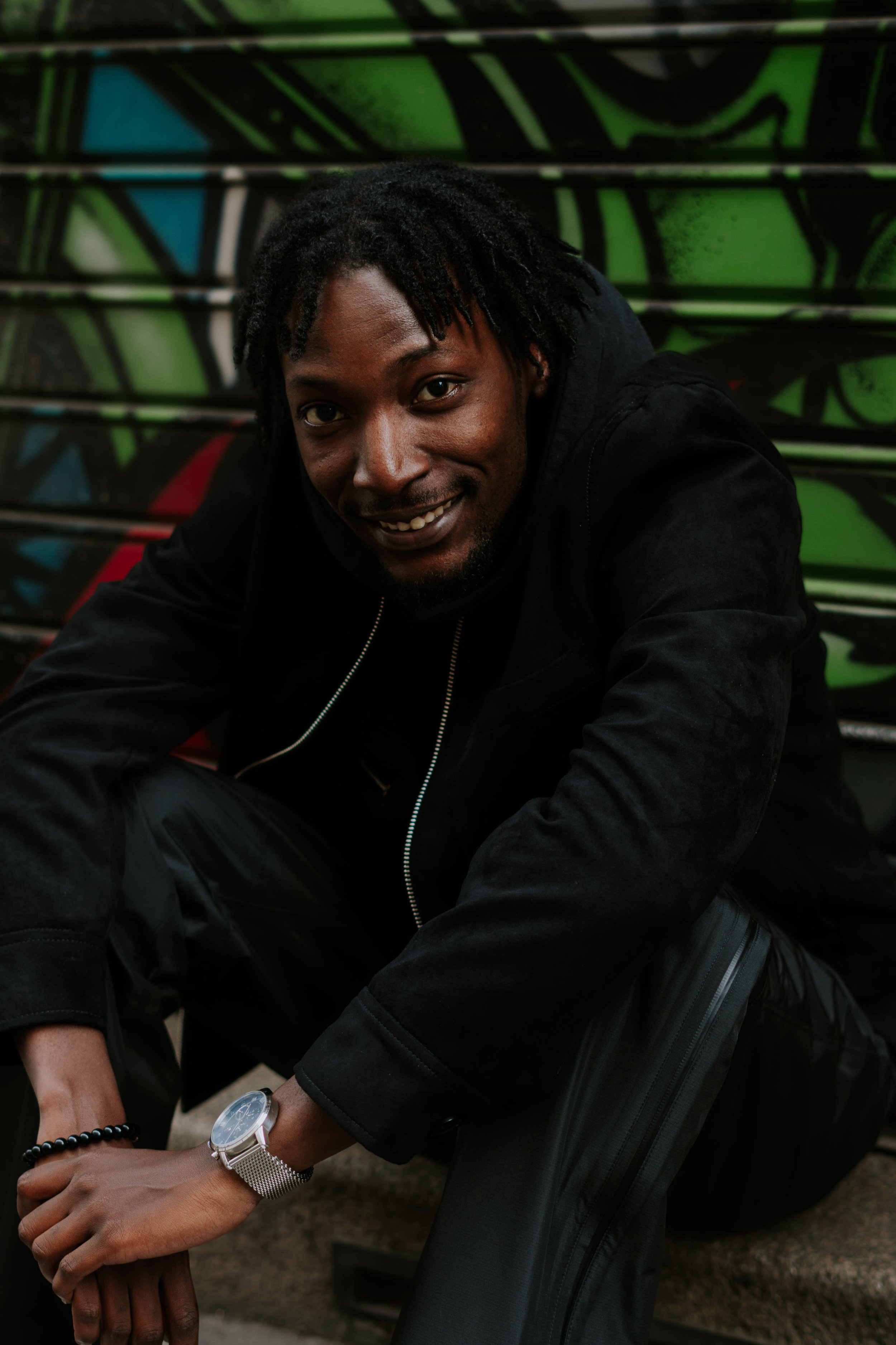 Un homme souriant avec des dreadlocks, portant une veste noire, un bracelet et une montre, assis devant un mur graffité coloré.