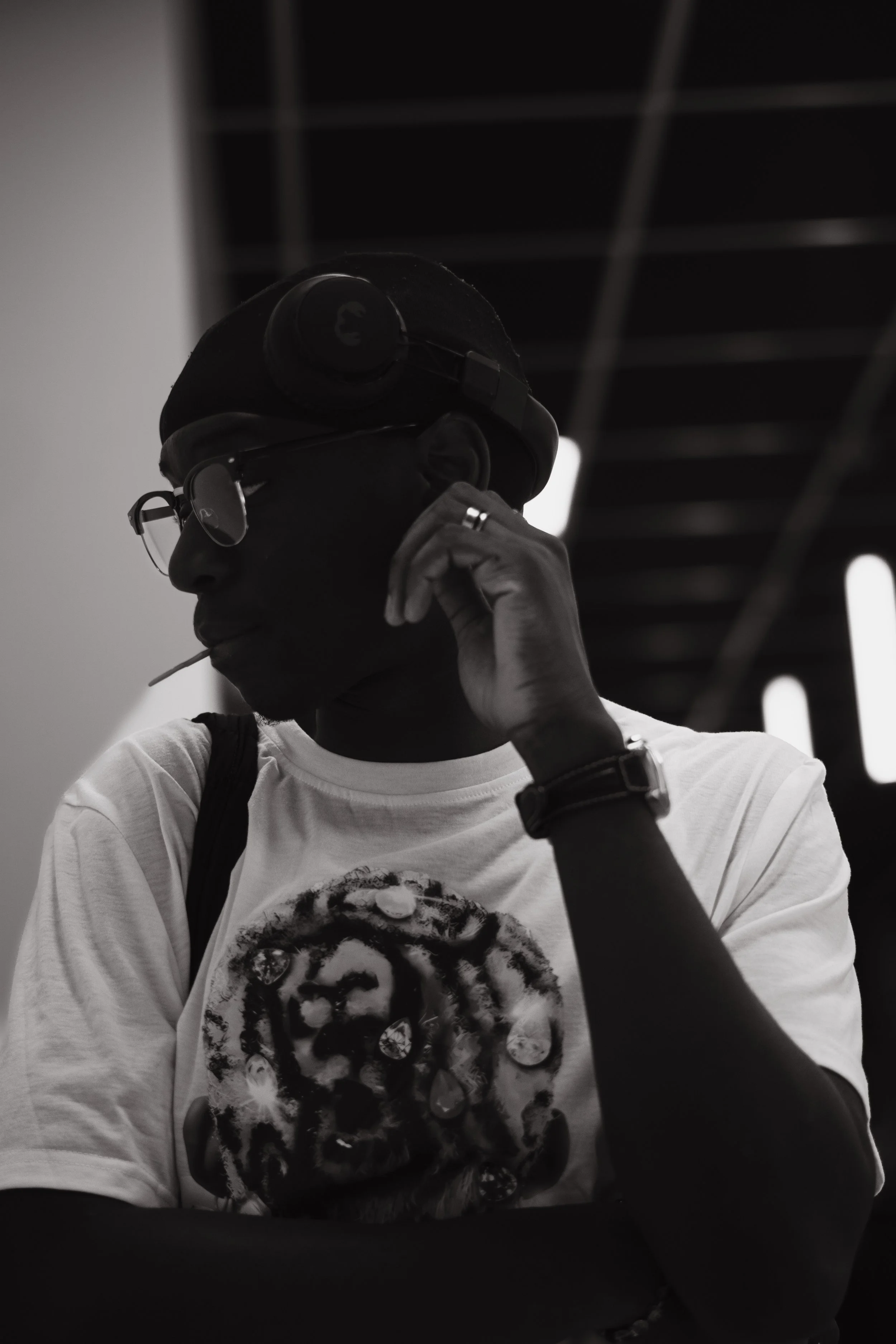 Un homme portant un casque de réalité virtuelle, des lunettes et une chemise blanche avec une image d'une tête de lion. Il tient un bâtonnet dans la bouche et porte une montre. L'arrière-plan est sombre avec des lumières verticales.