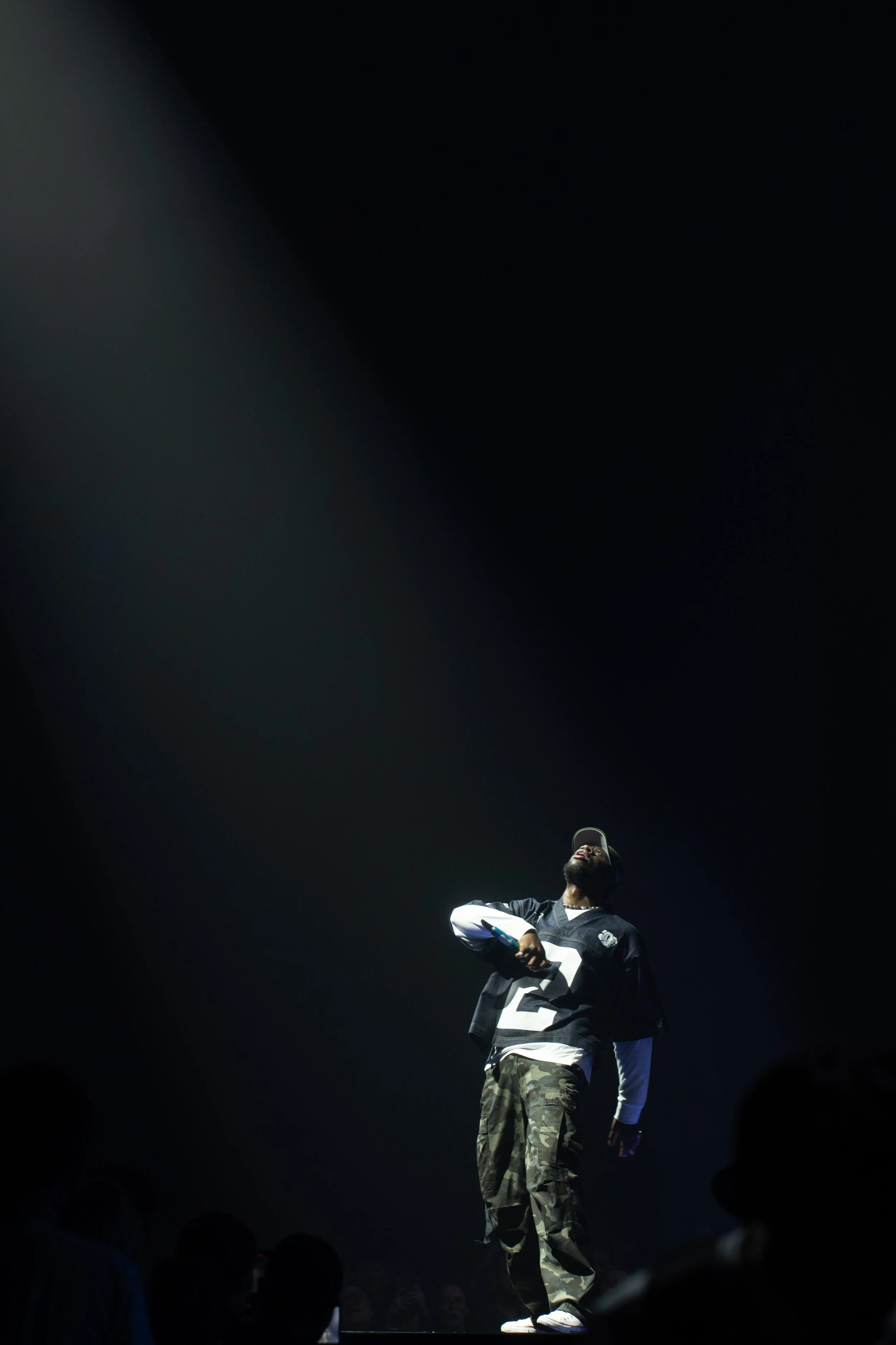 Un homme sur scène, éclairé par un faisceau de lumière, portant un maillot de football avec le numéro 2, des pantalons camouflage et une casquette, entouré d'un public.