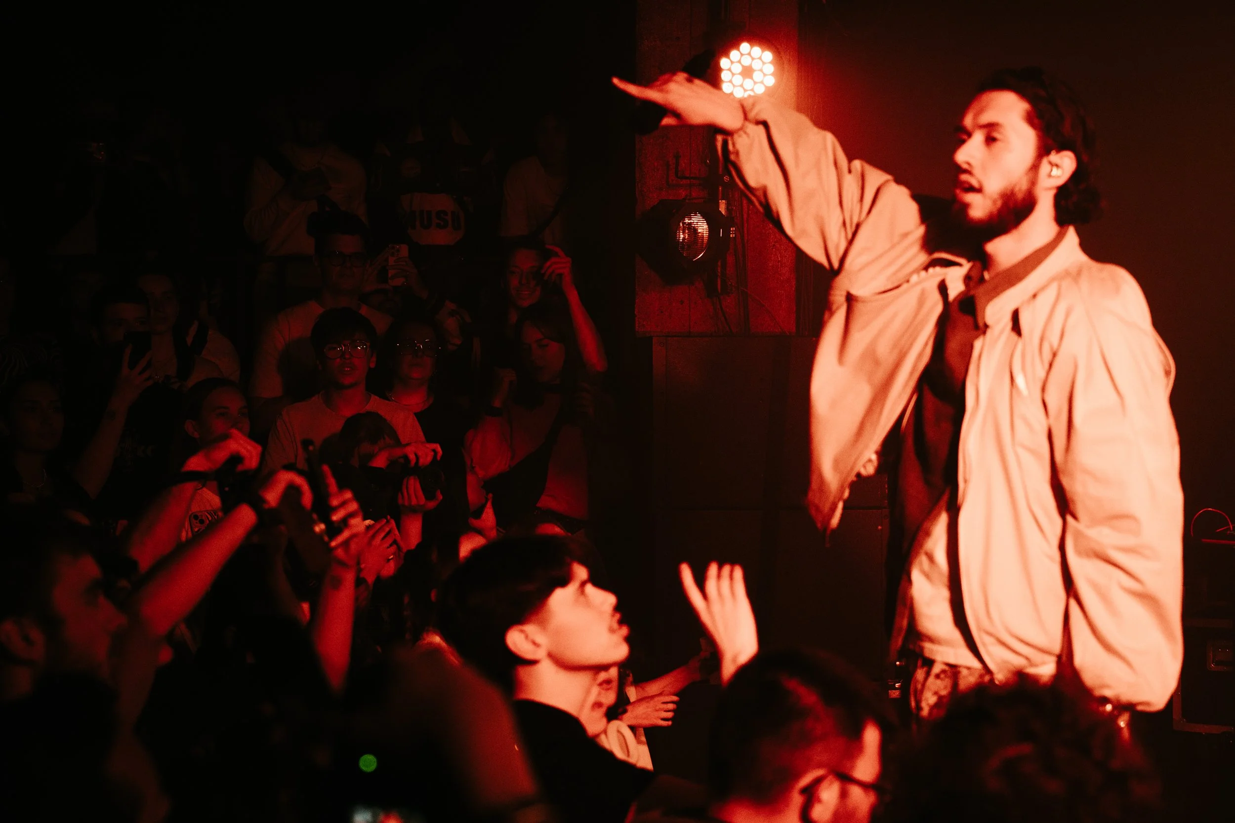 Une personne avec une barbe, portant une veste beige, s'adresse à un public lors d'une performance ou d'une conversation, dans un environnement sombre avec un éclairage rouge