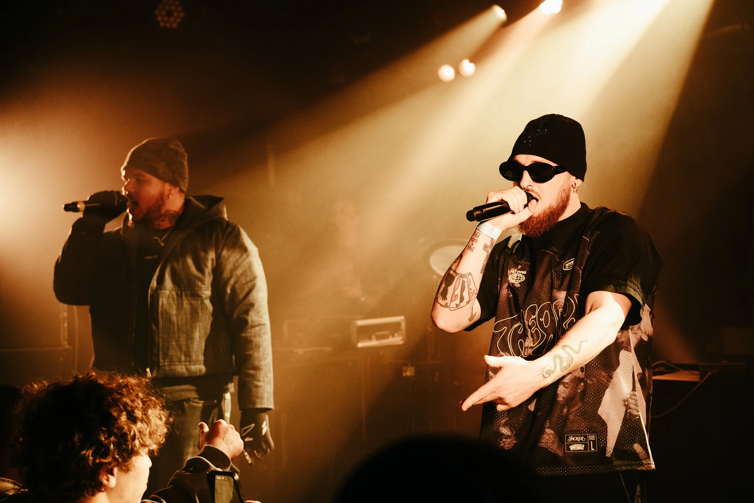 Deux rappeurs sur scène sous des lumières chaudes, l'un en veste en cuir et bonnet, l'autre en t-shirt à motifs, lunettes noires et bonnet, en train de performer. Audience en premier plan.
