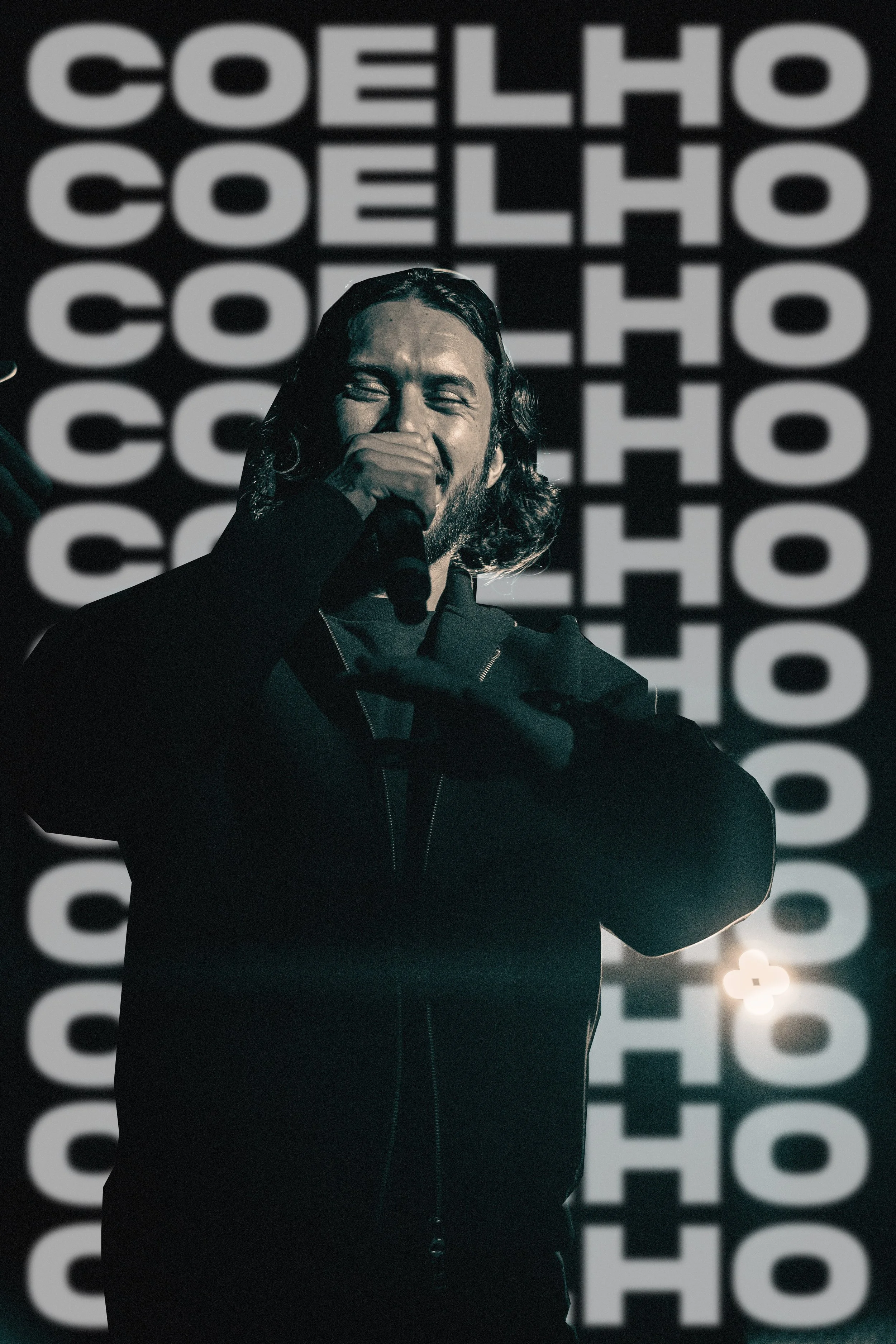 Un homme avec des cheveux longs et une barbe, tenant un microphone et souriant, se tient devant un fond avec le mot 'COELHO' répété en blanc sur fond noir.