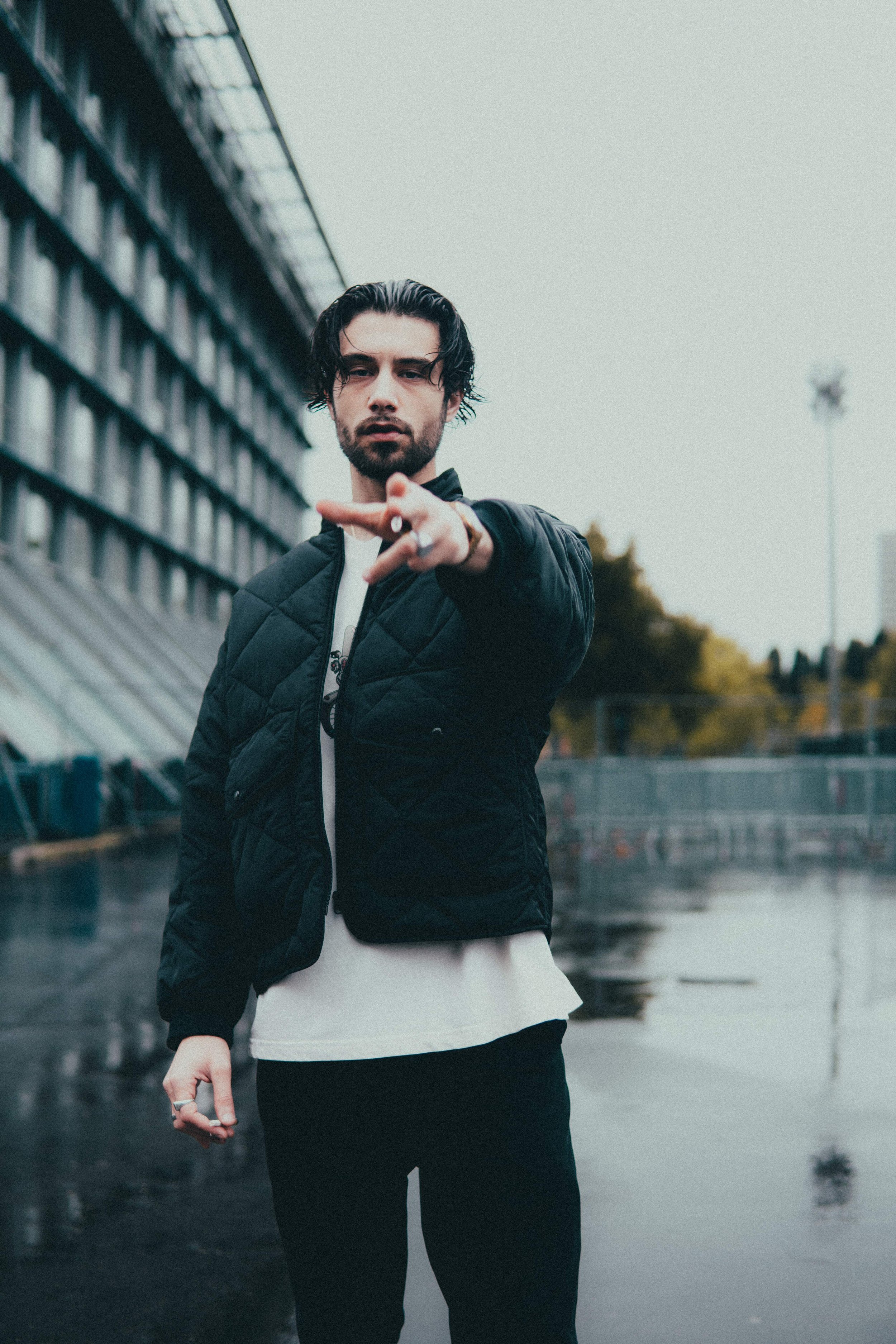 Jeune homme avec des cheveux longs et une barbe, portant une veste noire, faisant un geste de la main vers la caméra, dans un environnement urbain avec un bâtiment en arrière-plan et une surface mouillée.