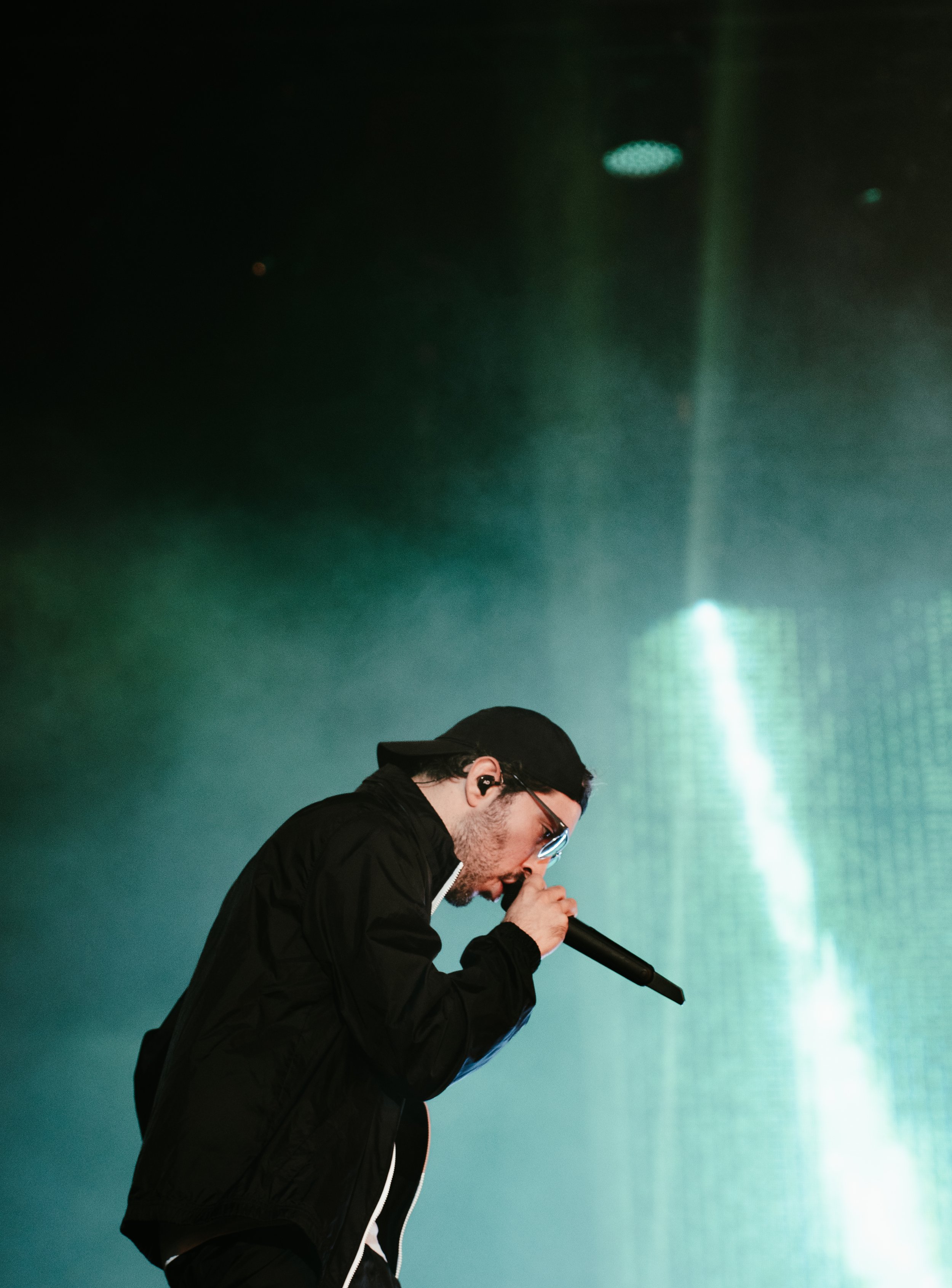 Un homme chante en tenant un microphone sur scène, vêtu d'une veste noire, portant des lunettes de soleil et un bandeau noir, avec un éclairage de scène vert et bleu en arrière-plan.