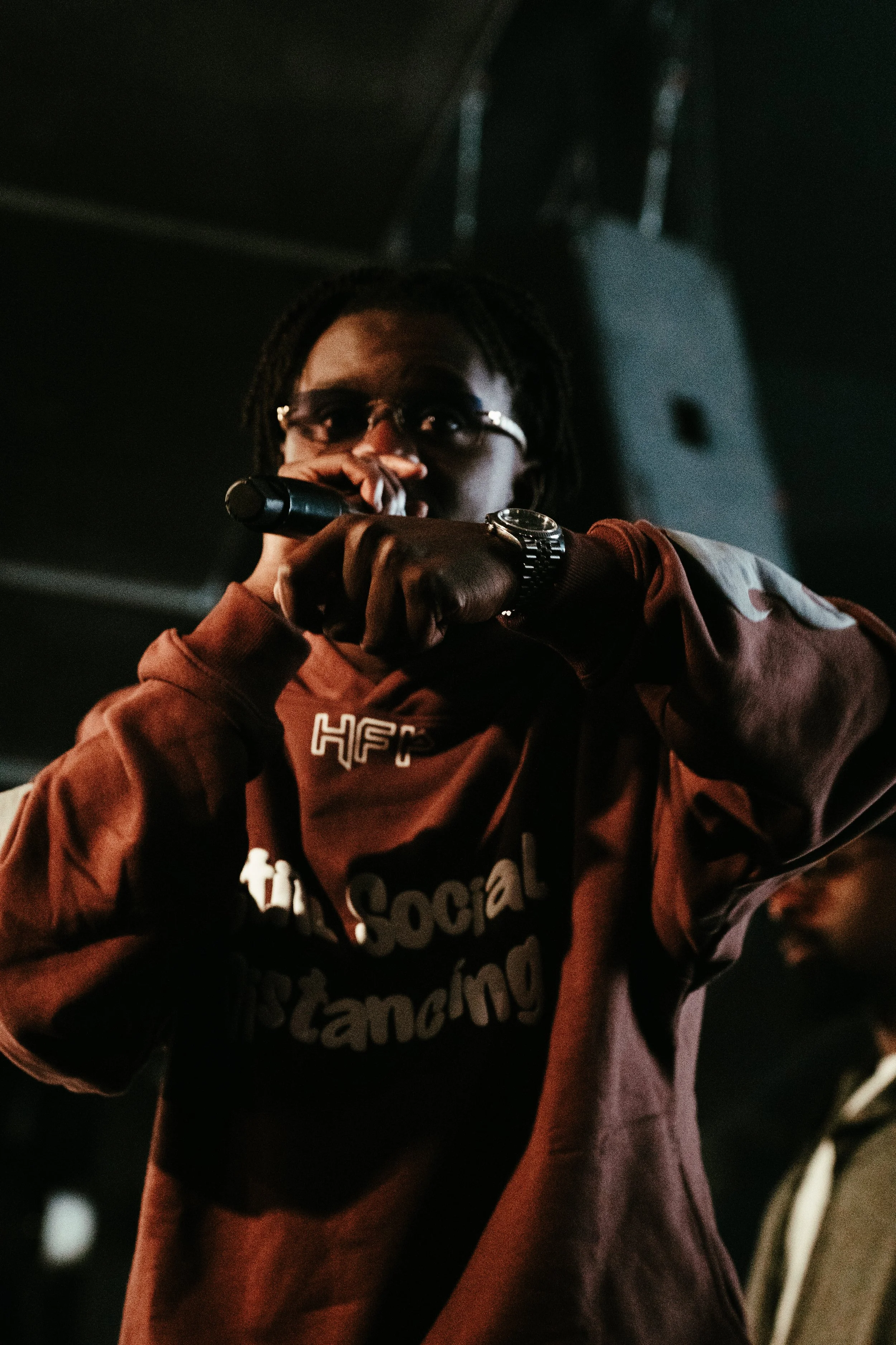 Un jeune homme noir portant des lunettes, un sweat à capuche marron avec des inscriptions, tient un microphone et semble en train de chanter ou de parler lors d'une performance ou d'un événement.