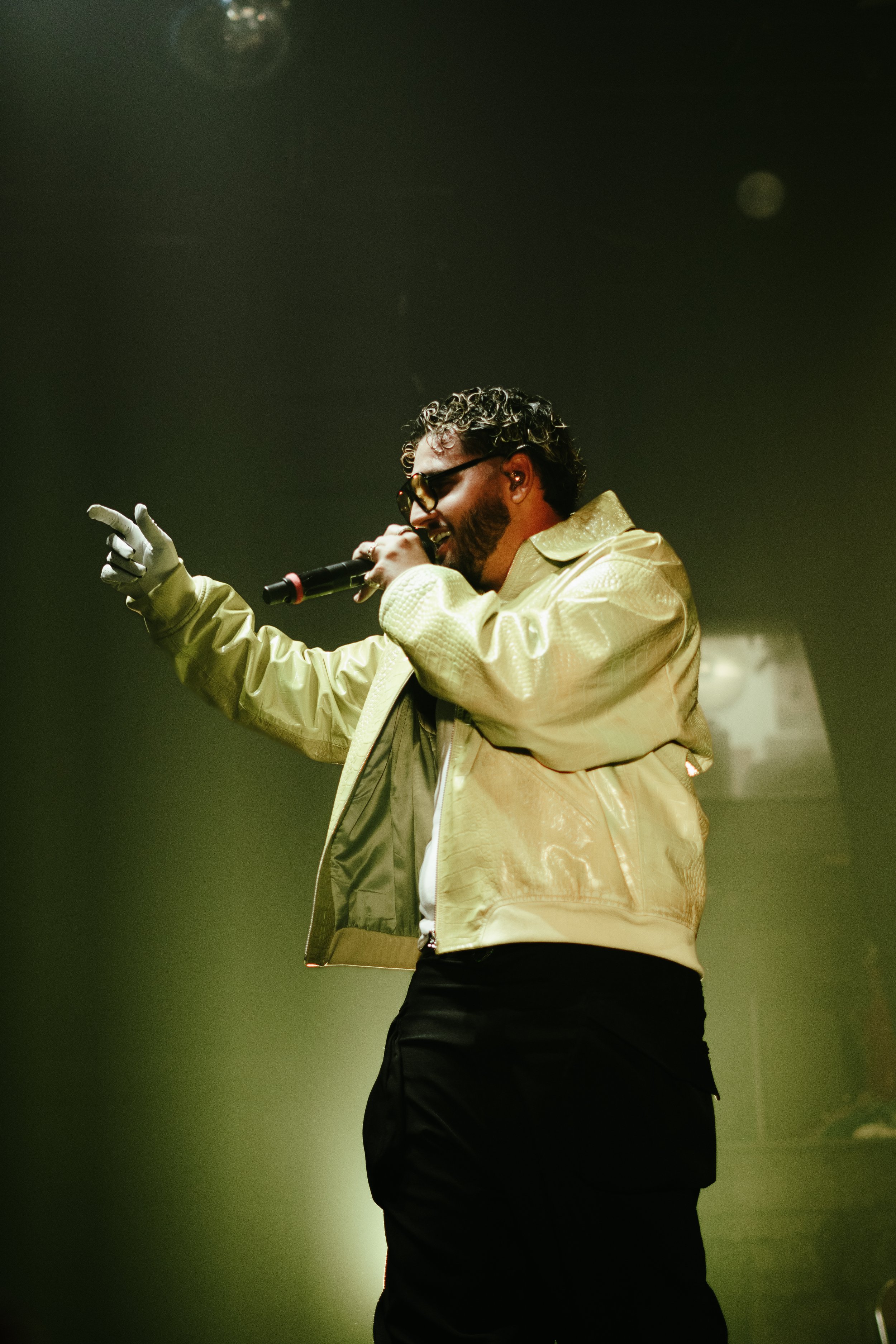 Un homme chante sur scène, portant des lunettes de soleil, une veste beige brillante, et tenant un microphone dans la main.