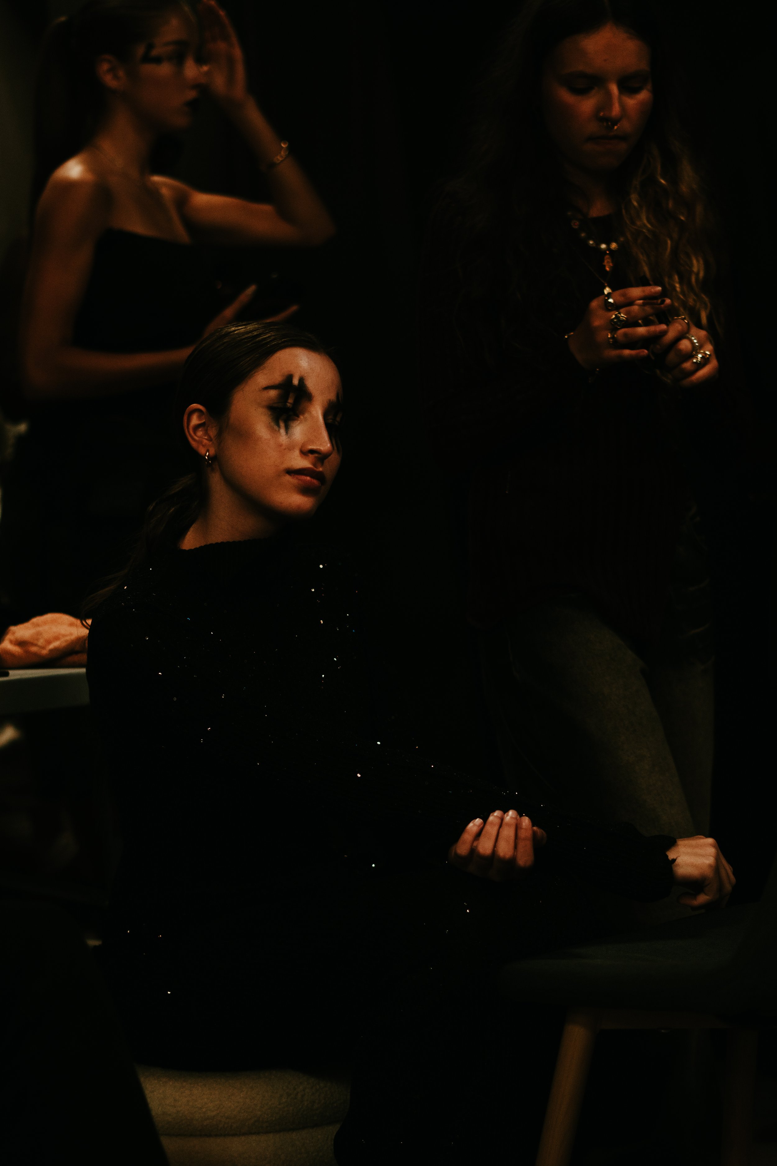 Une jeune femme assise dans une pièce sombre, portant une robe noire à paillettes, avec un maquillage artistique autour des yeux, une autre femme debout à côté, et une troisième en arrière, toutes dans une atmosphère sombre et mystérieuse.