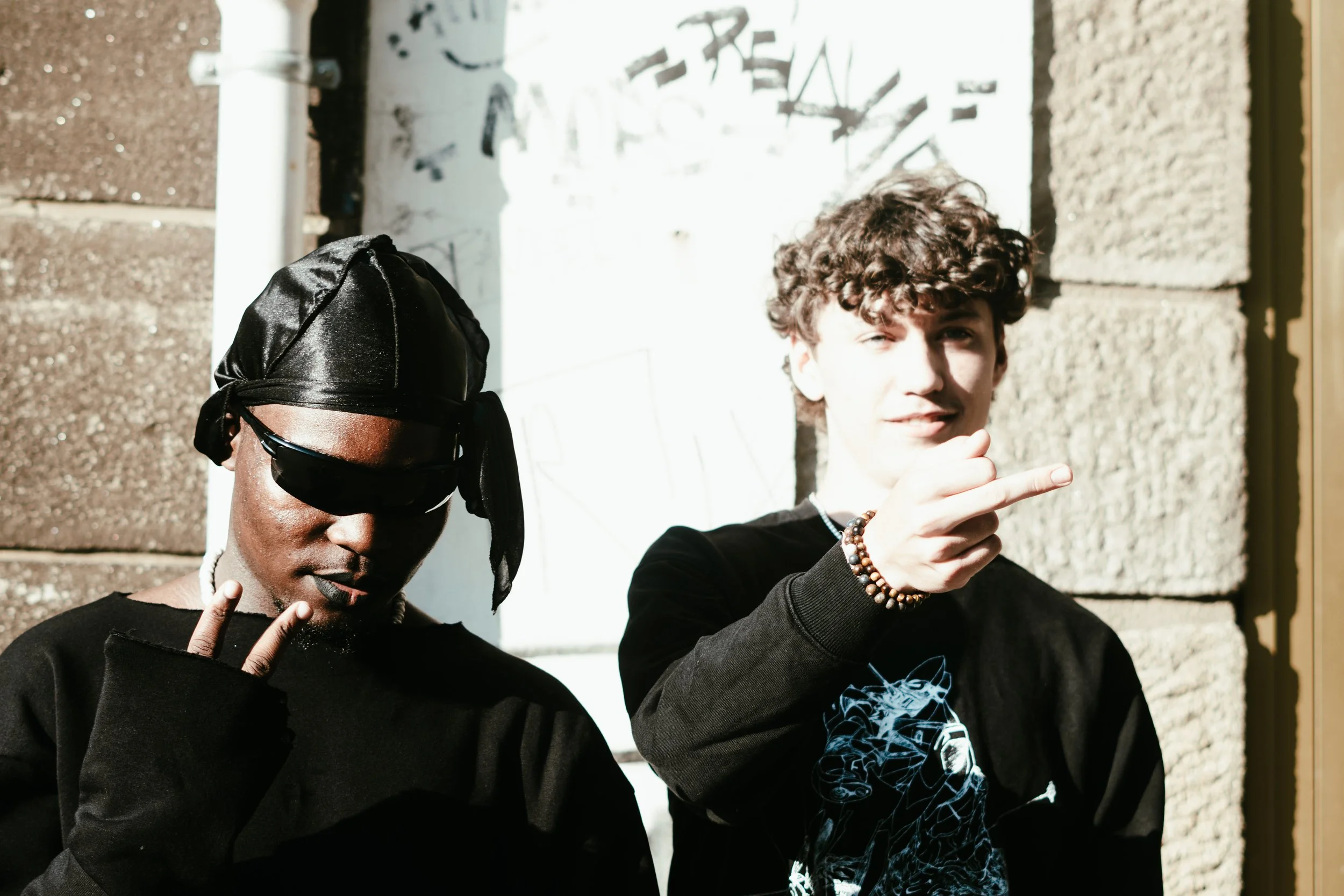 Deux jeunes hommes posent devant un mur en pierre, l'un porte un bandeau noir et des lunettes de soleil, l'autre fait un geste de la main.