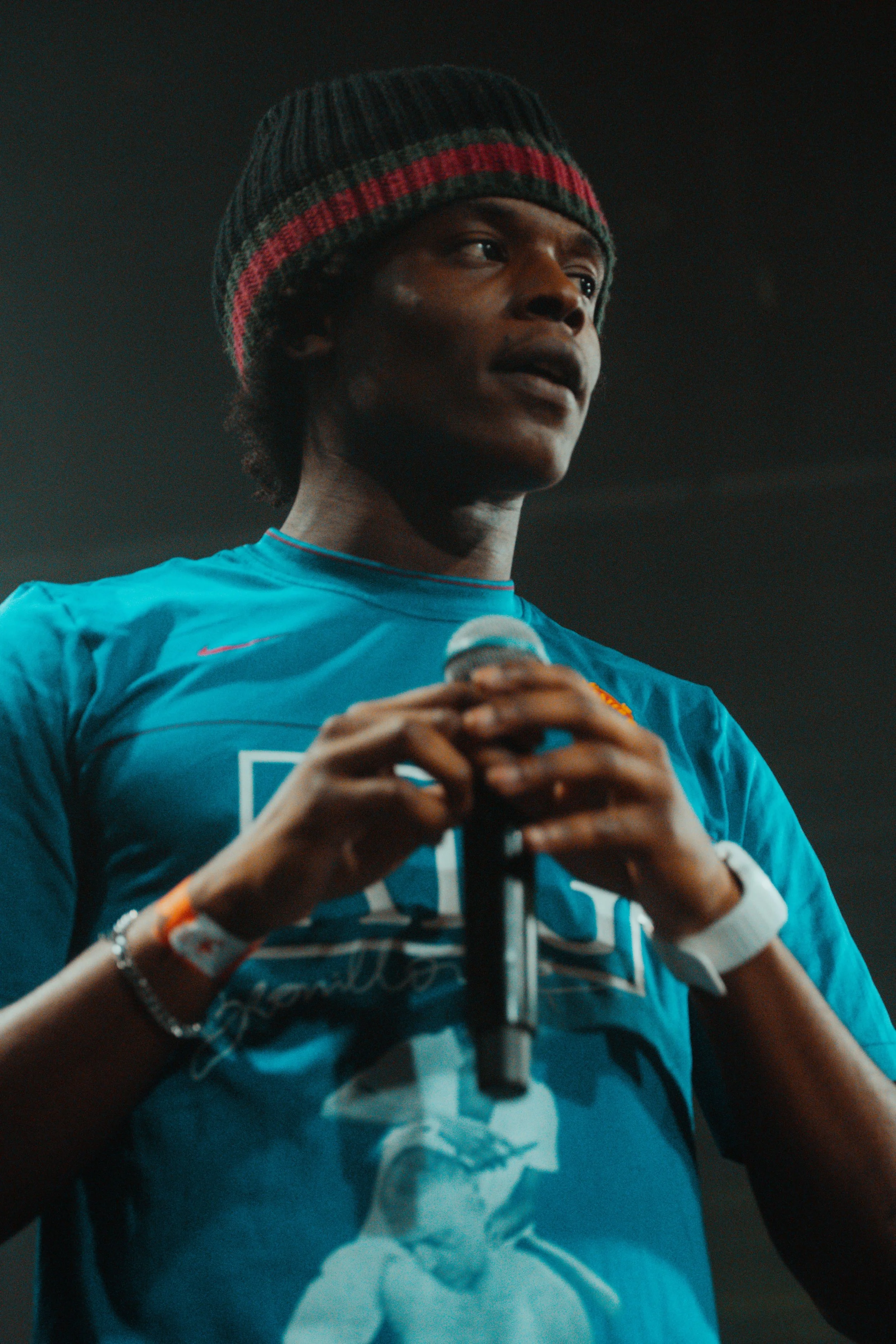 Un homme noir portant un bonnet et un t-shirt bleu, tenant un microphone, lors d'une performance ou d'une présentation.