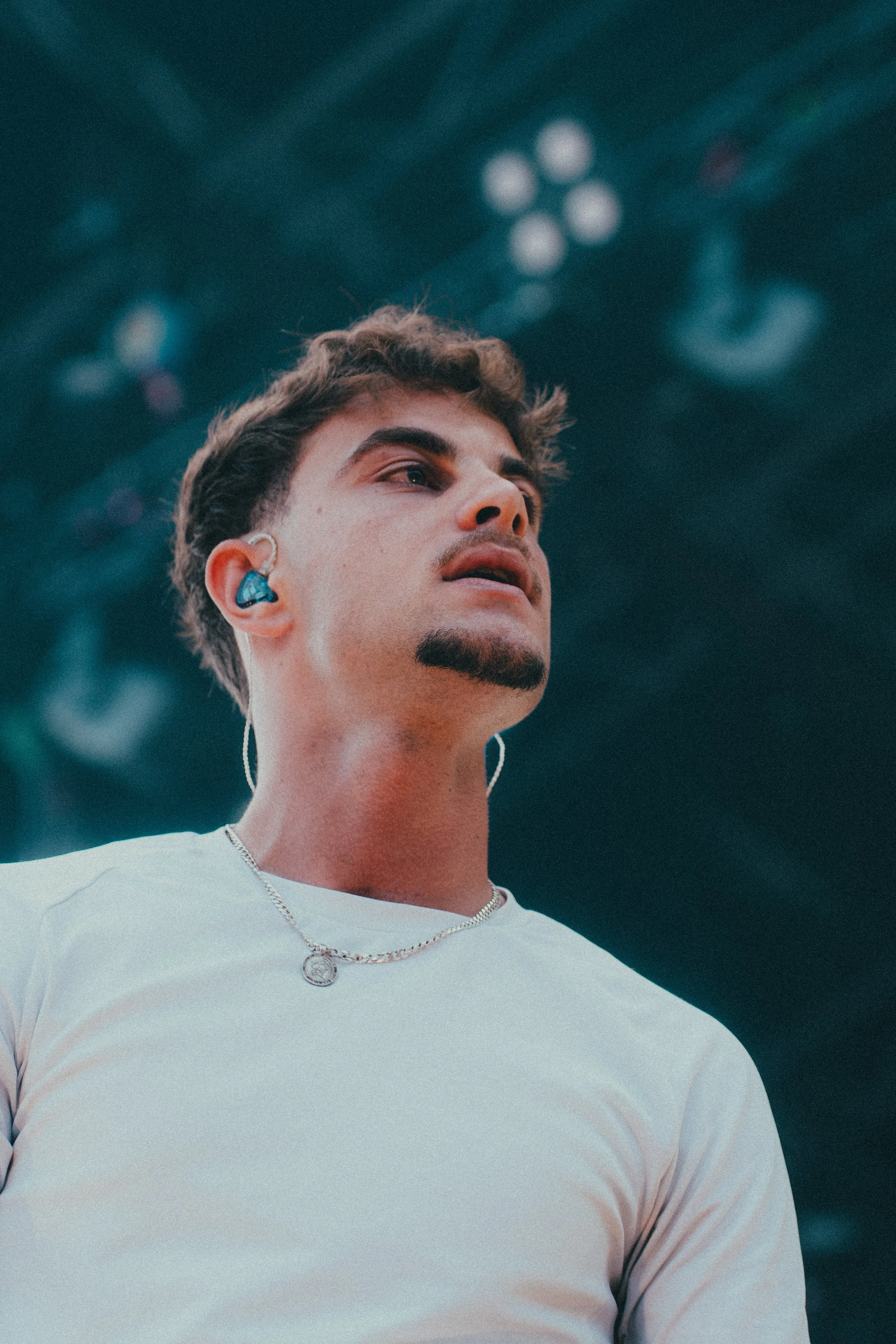 Un homme jeune avec des écouteurs bleus, portant un t-shirt blanc et une chaîne en argent, lors d'un concert ou d'un événement sur scène.