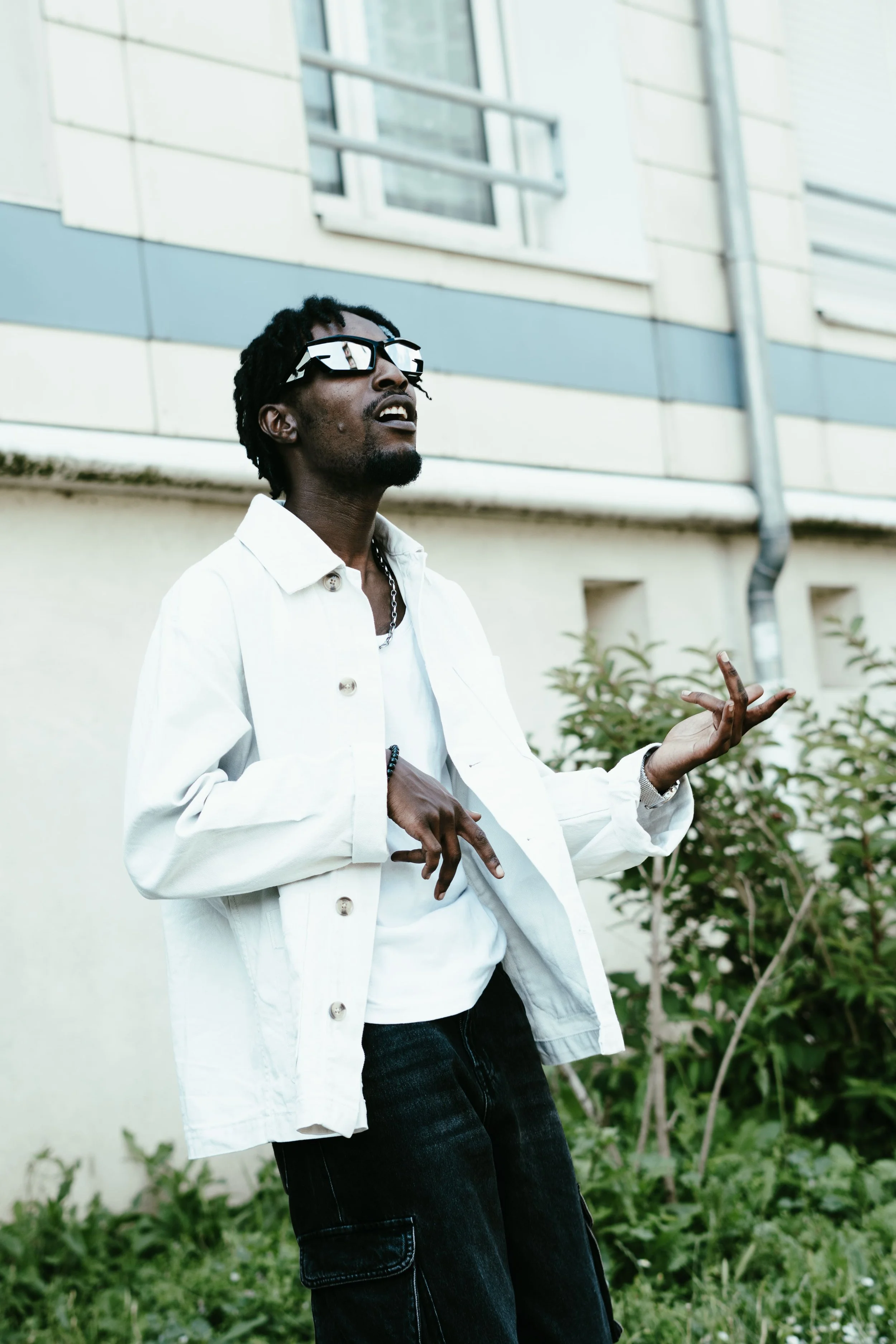 Un homme portant des lunettes de soleil, une chemise blanche, un t-shirt blanc, et un pantalon noir, se tenant à l'extérieur devant un bâtiment avec des fenêtres et des plantes
